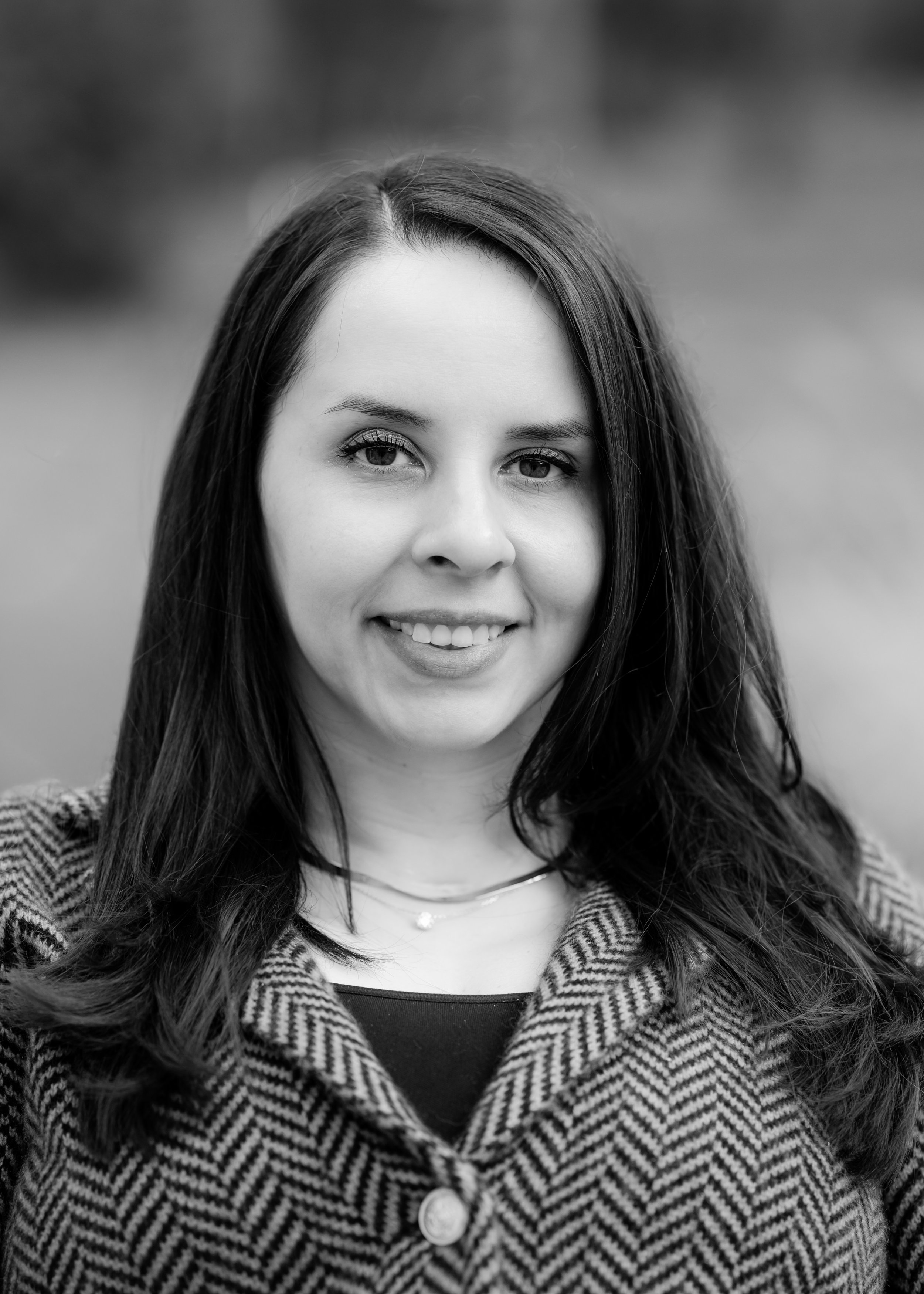 Tierney Cauley, owner of Eunoia Mental Health and Wellness in NJ, with long dark hair smiling, wearing a striped coat and a top with a necklace, outdoors with a blurred background.