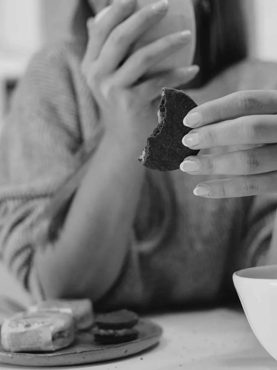 A person holding a bitten cookie with one hand, with a plate of cookies and a bowl on the table in front.