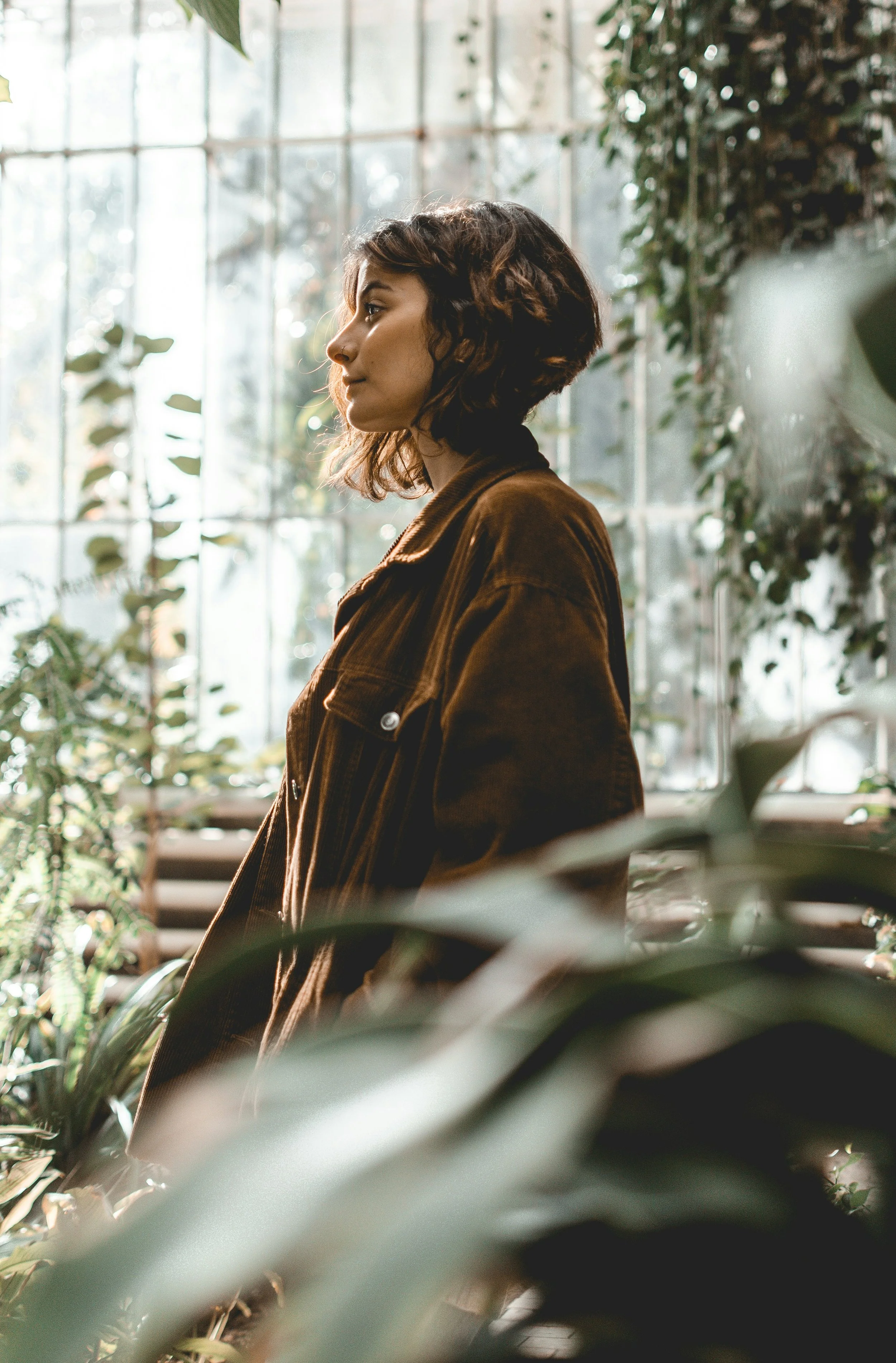 A woman with short, wavy hair wearing a brown jacket, standing in a greenhouse or botanical garden with sunlight streaming through large windows, surrounded by green plants and foliage.