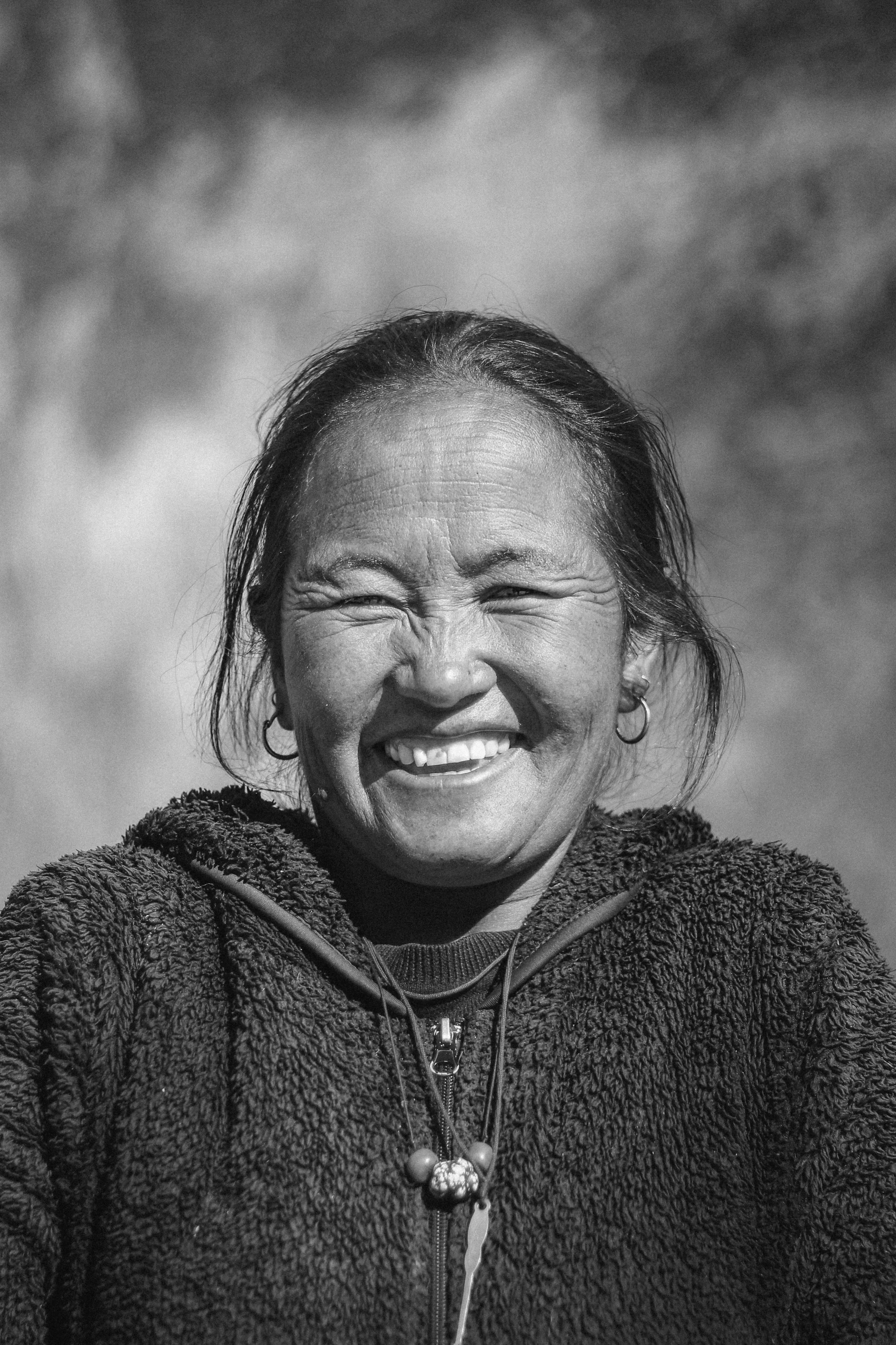 A black and white photo of a smiling woman with her eyes closed, wearing a zip-up fleece jacket, earrings, and a necklace, outdoors with a cloudy sky in the background.