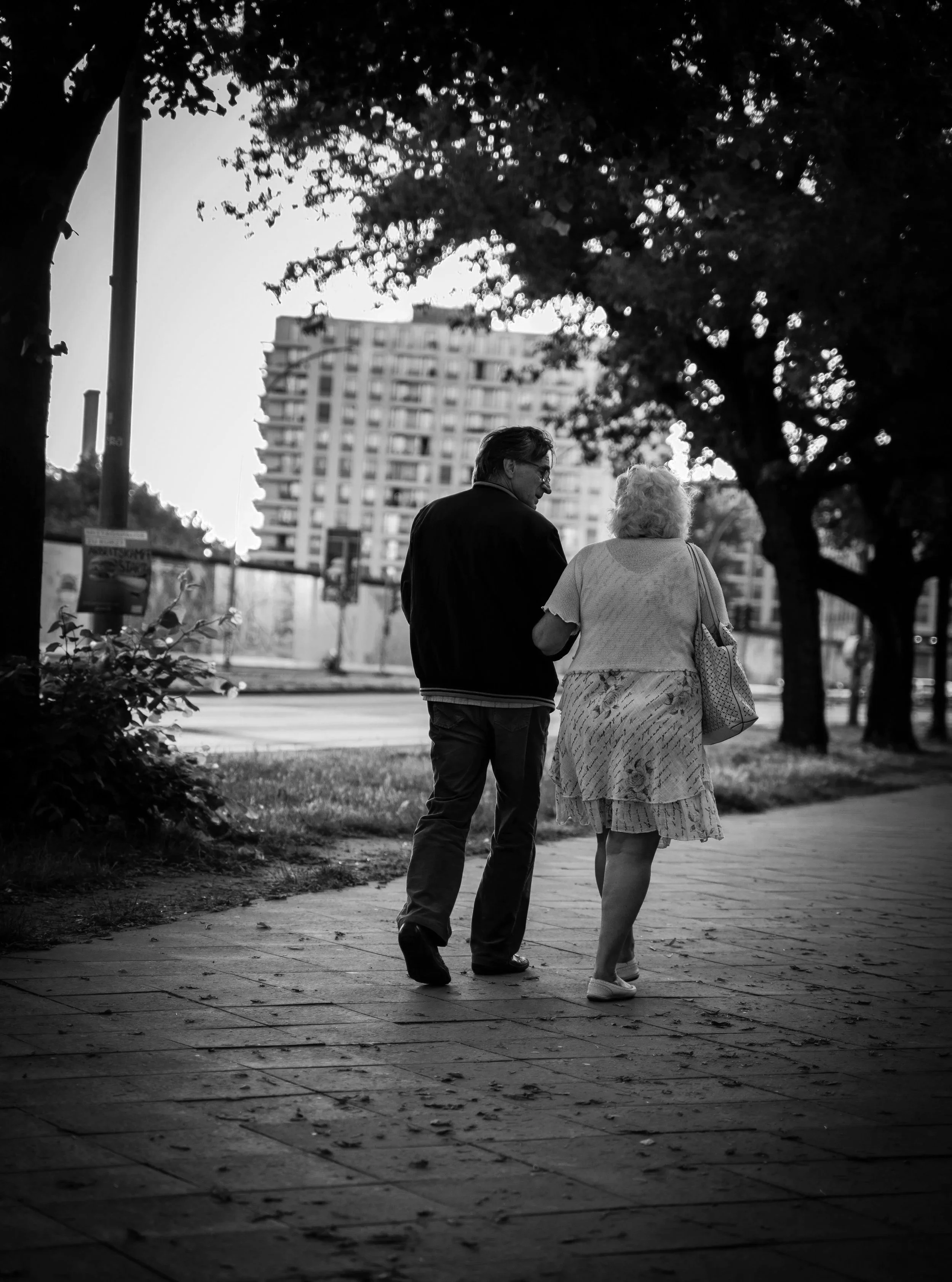 A black-and-white photo of an elderly woman and a middle-aged man walking together on a city sidewalk, surrounded by trees and buildings.