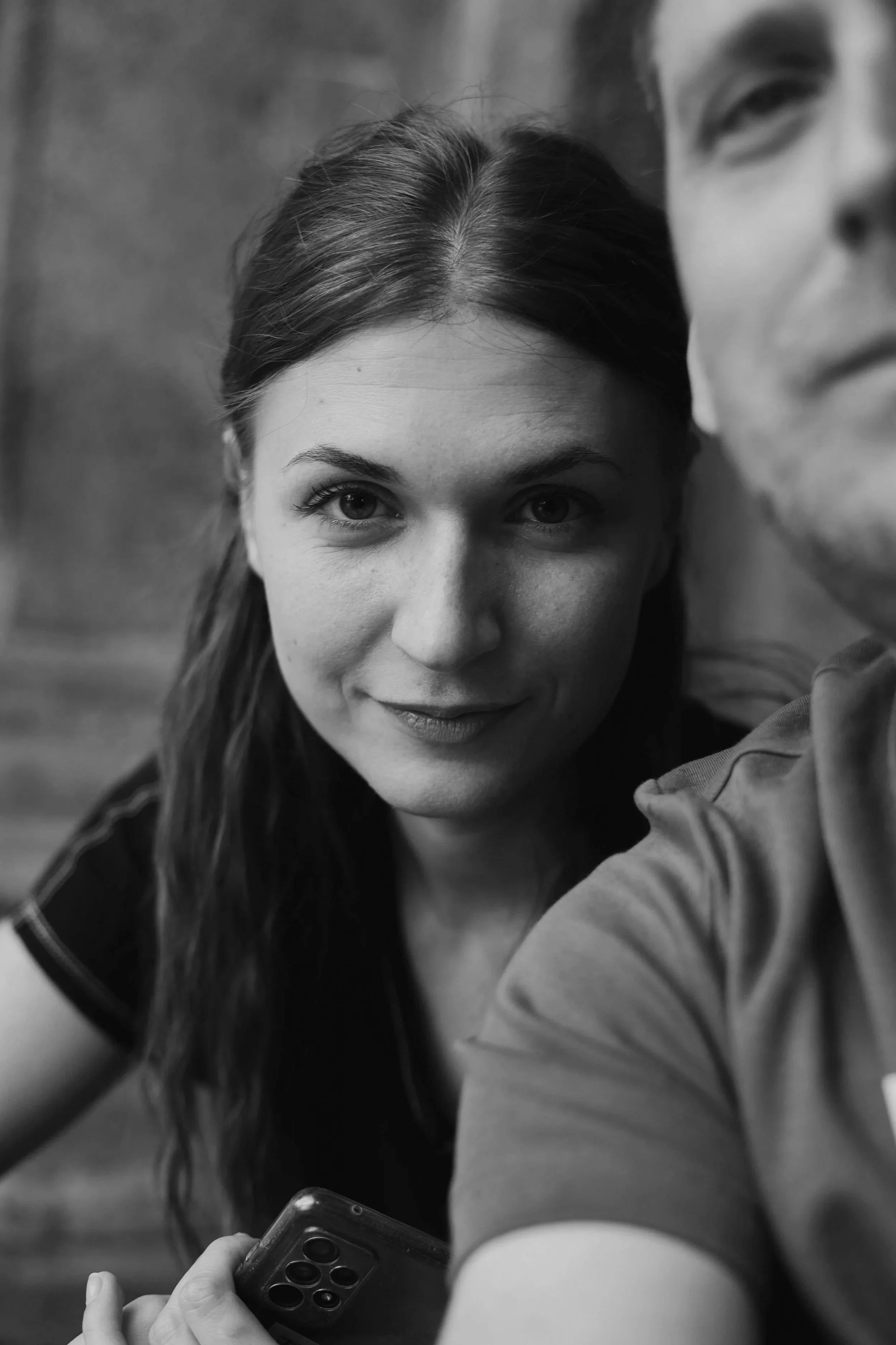 A black-and-white photo of a young woman with long hair holding a smartphone, partially obscured by a person's arm in the foreground. She is smiling and looking at the camera.