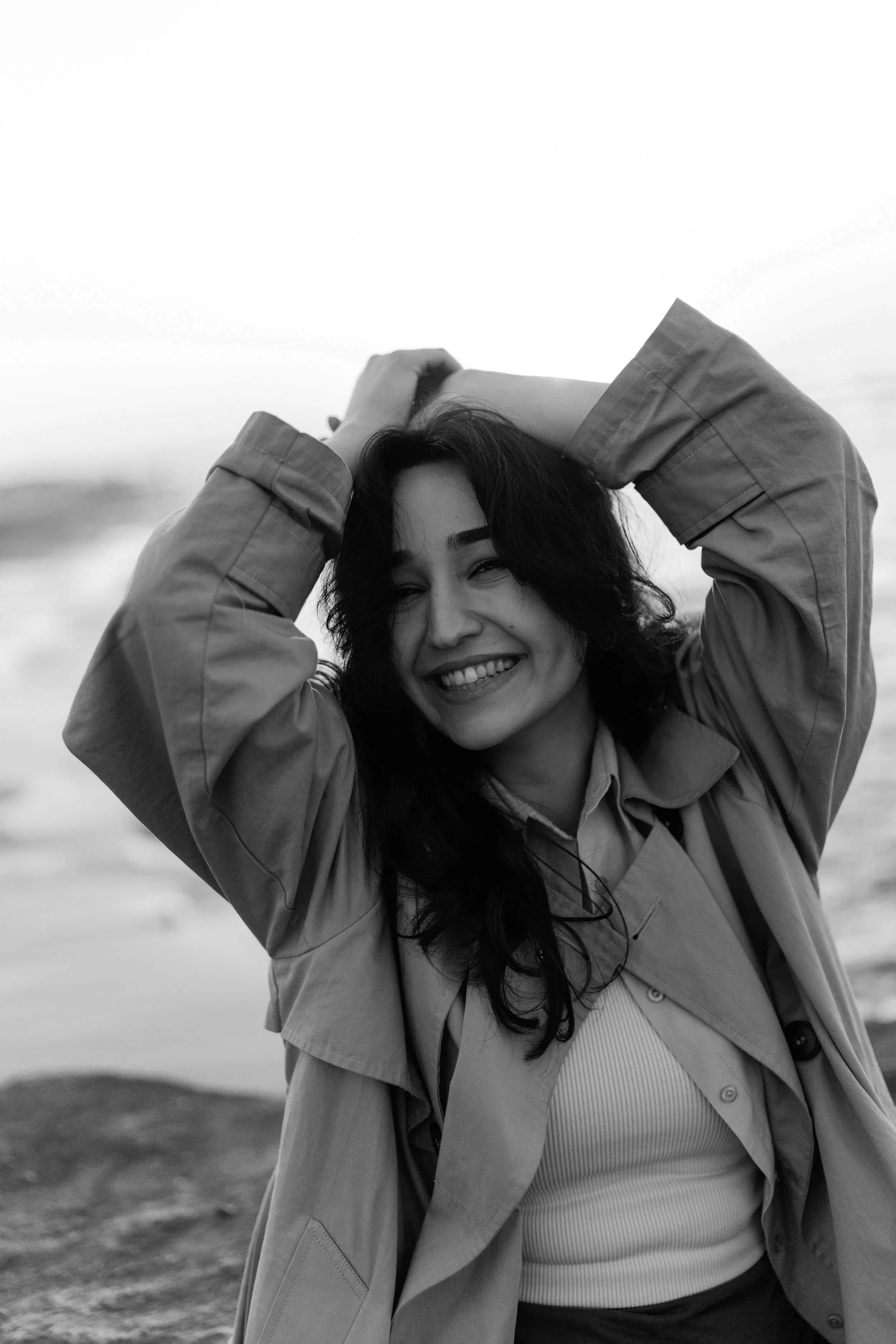 A woman smiling with her hands behind her head, wearing a coat and a striped shirt, standing outdoors near water in black and white.