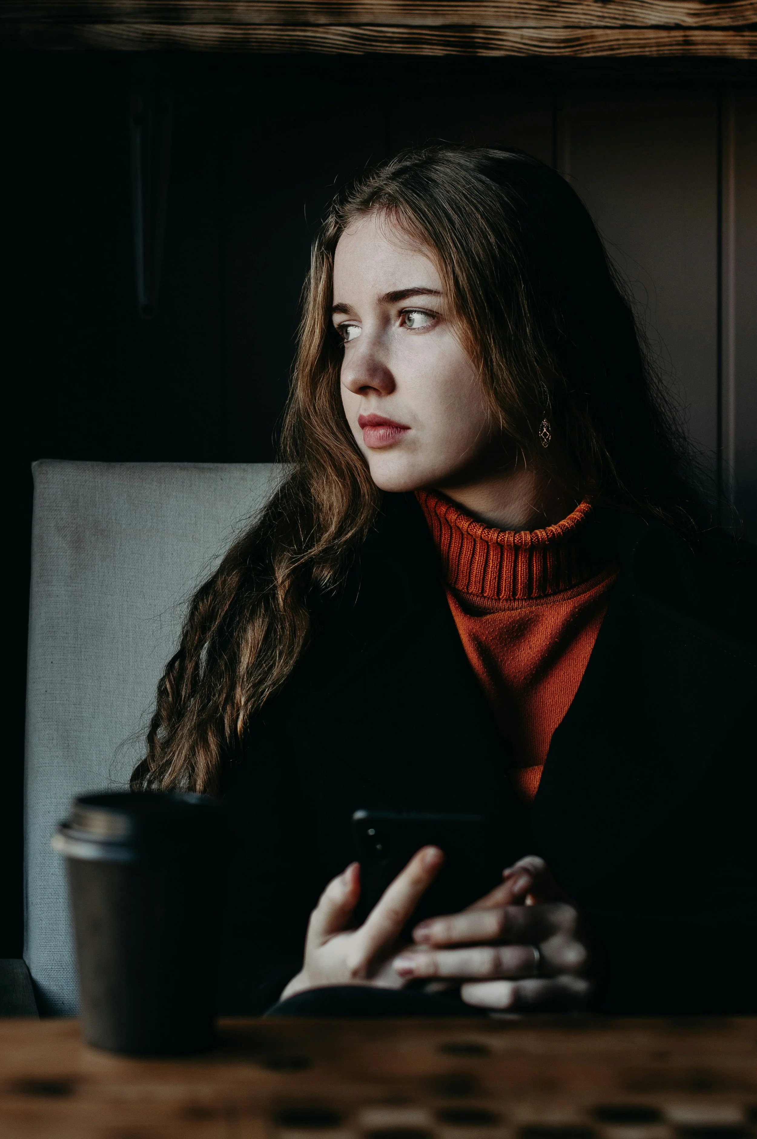 A young woman with long brown hair and light skin sitting at a table, looking to her right while holding a smartphone, with a coffee cup on the table in front of her.