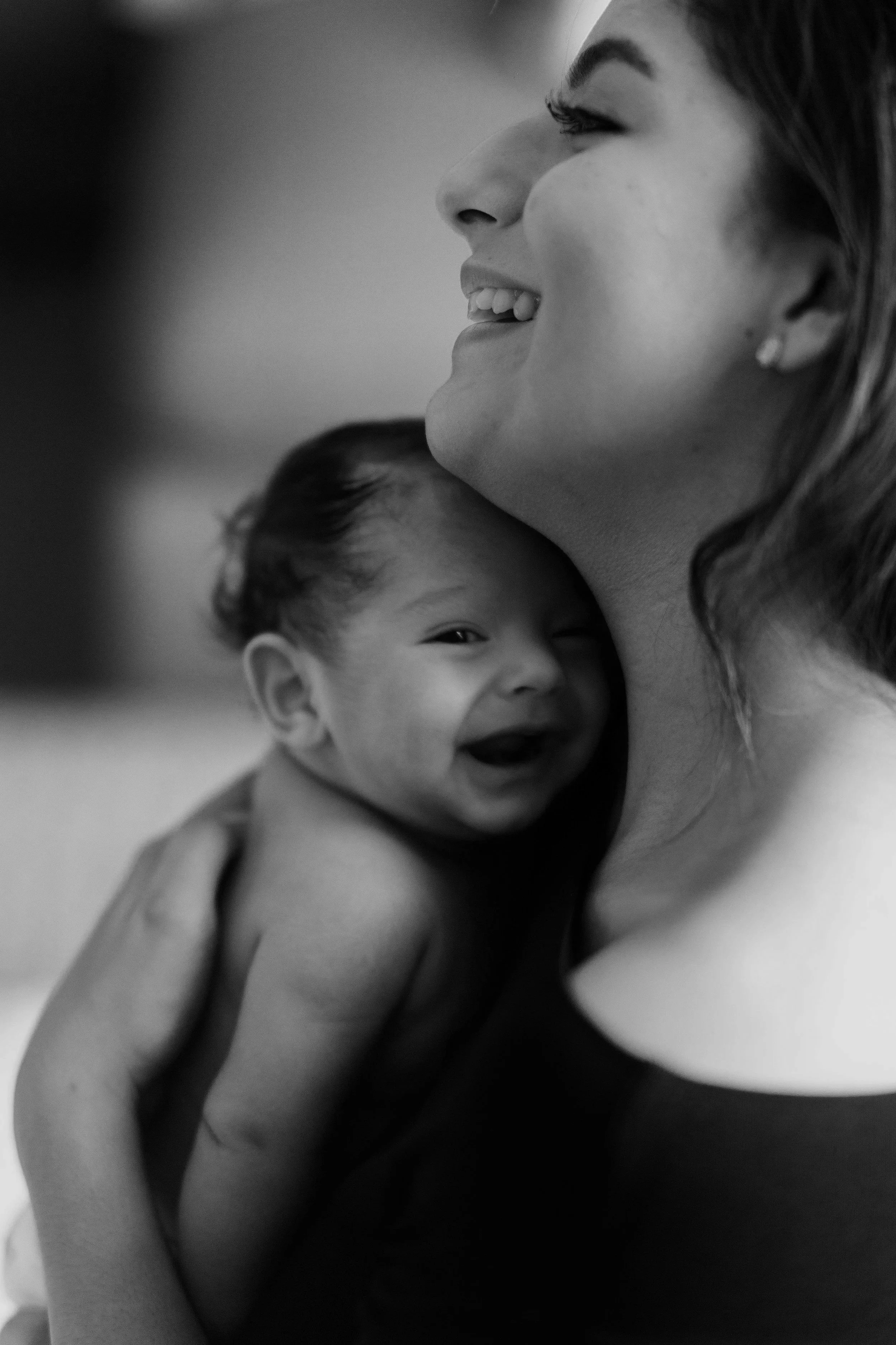 A woman with long hair and pearl earrings is smiling and embracing a young child, who is laughing and looking at the camera.