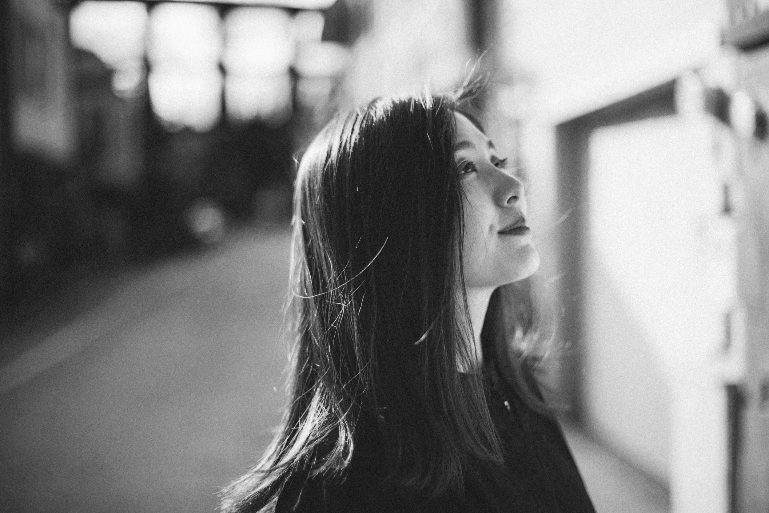 A black-and-white photo of a woman with long hair, looking upward in a hallway or corridor with lockers or cabinets on the side and blurred lights in the background.
