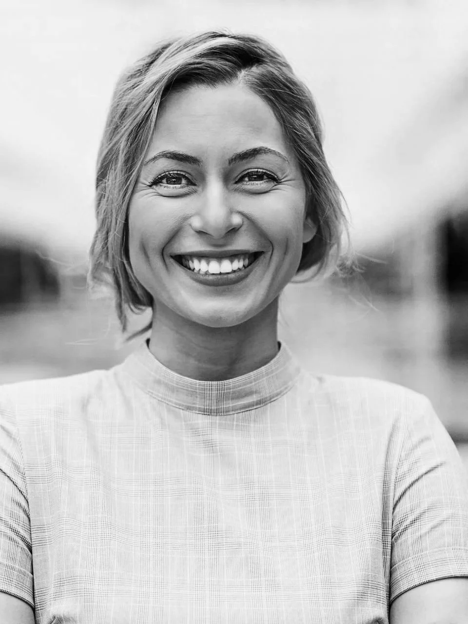 Black-and-white photo of a smiling woman with short, wavy hair wearing a high-neck shirt, outdoors.