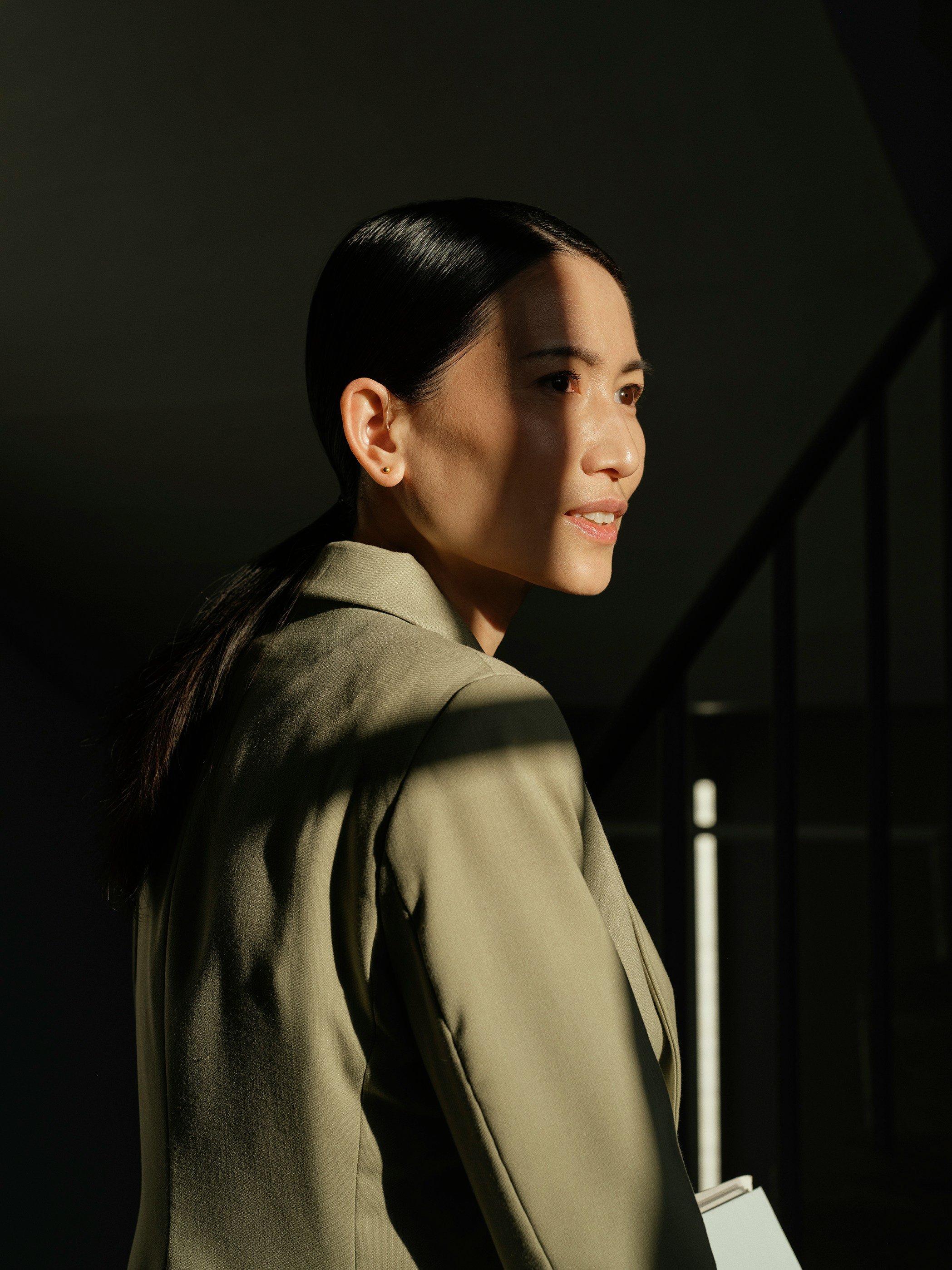 A woman with slicked-back black hair and earrings, wearing a beige blazer, looks to the side with a slight smile, against a dark background with a light source creating a shadow on her face.