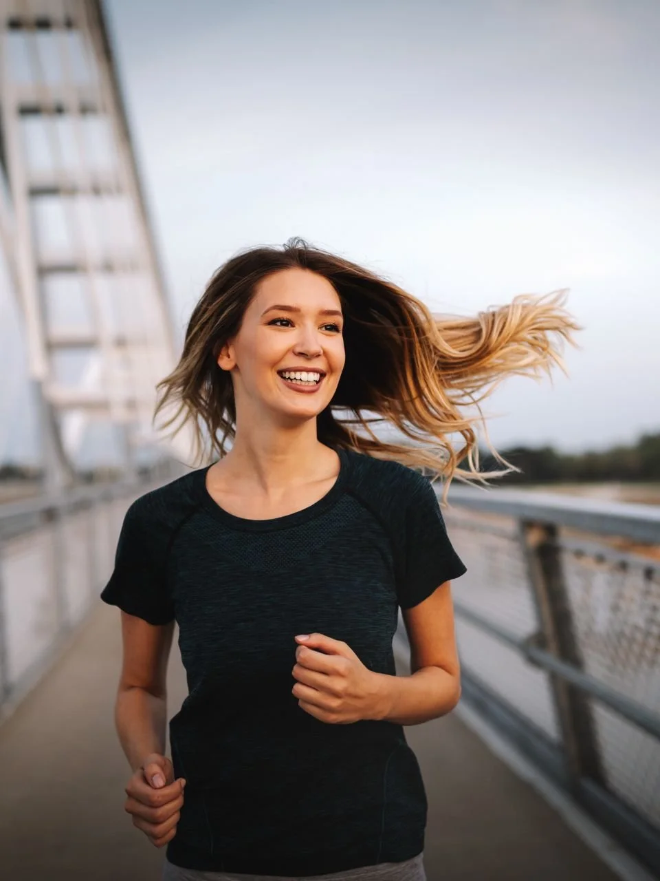 A smiling woman with long hair running on a bridge during daytime.