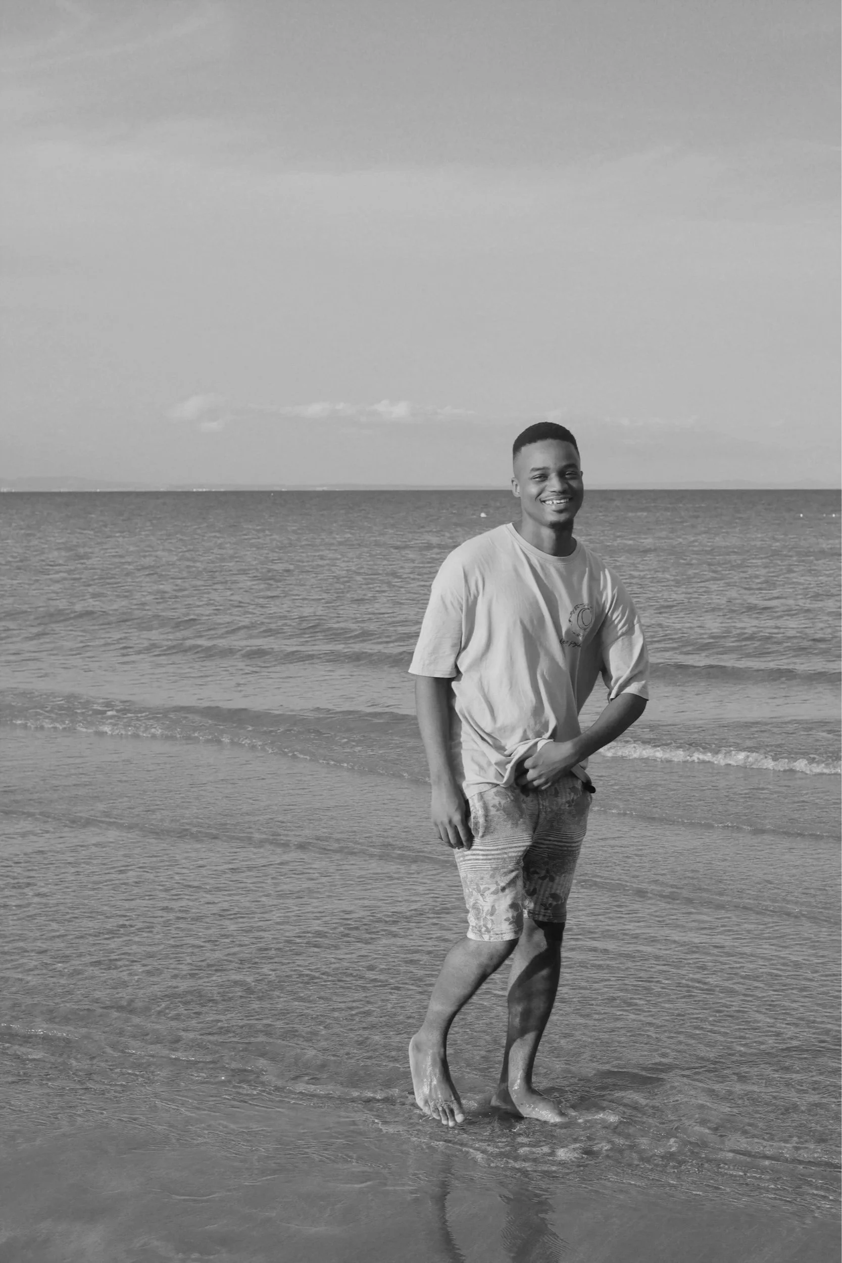 A smiling young man standing at the shoreline of an ocean or lake, with waves gently reaching his feet, under a partly cloudy sky.
