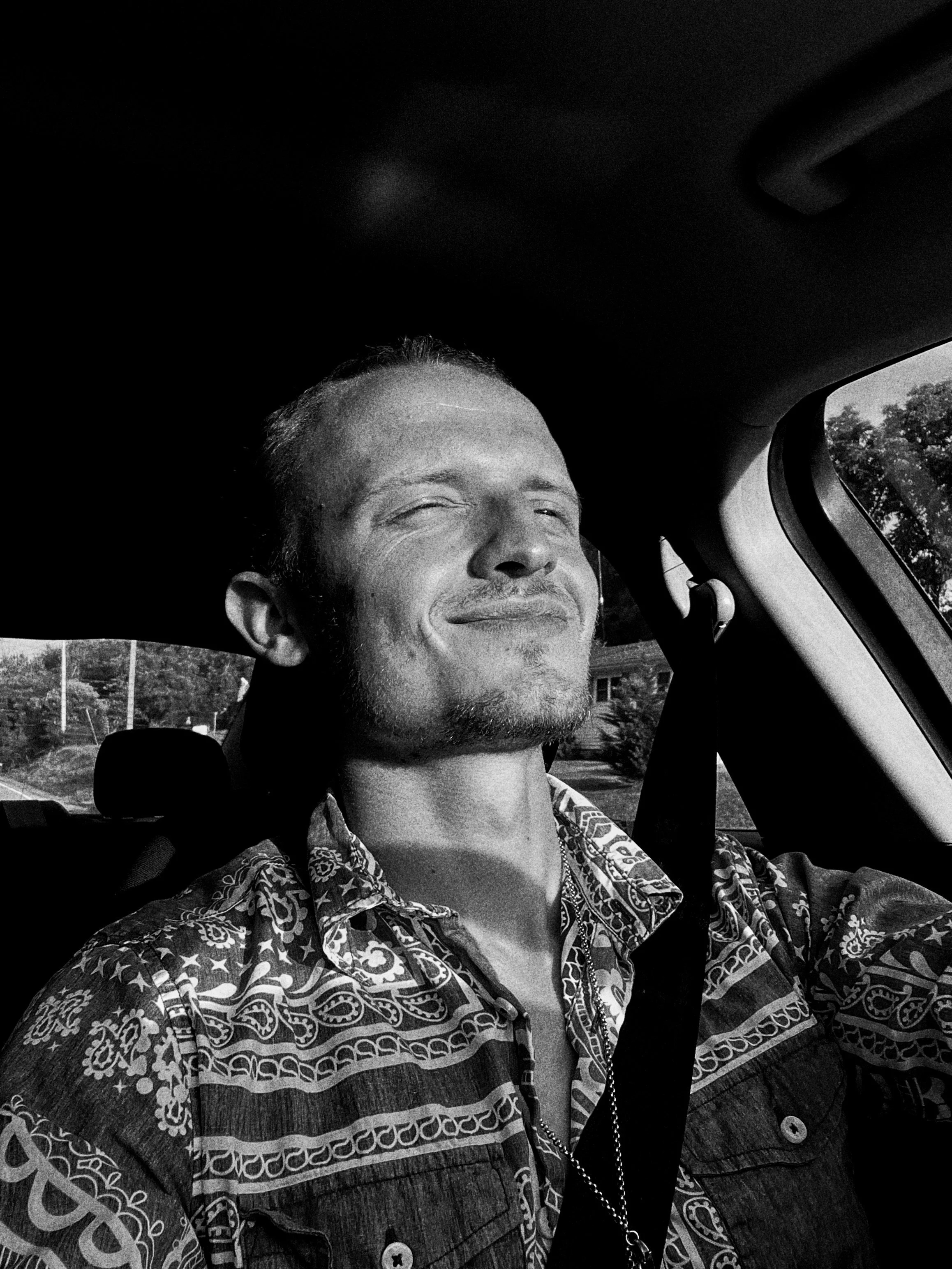 Black and white photo of a smiling man with closed eyes, wearing a patterned shirt, sitting inside a car with the door closed, with trees visible through the window.