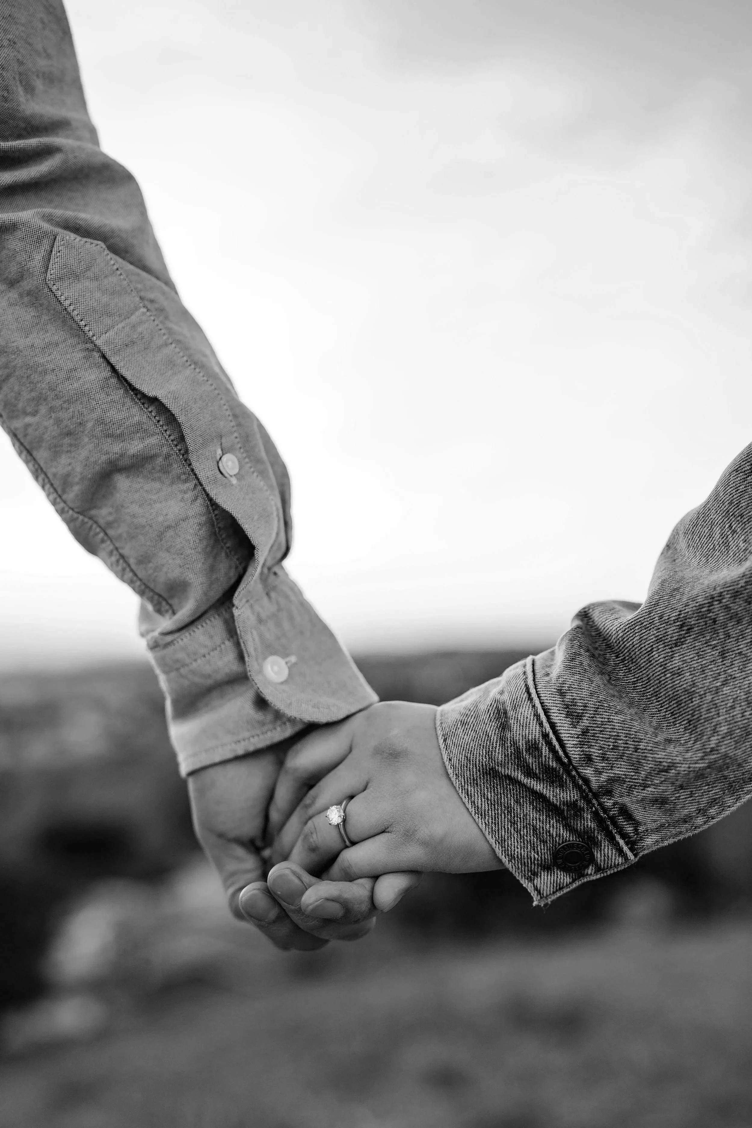 Close-up of two people holding hands, with one wearing a ring, outdoors with a blurred landscape in the background, in black and white.