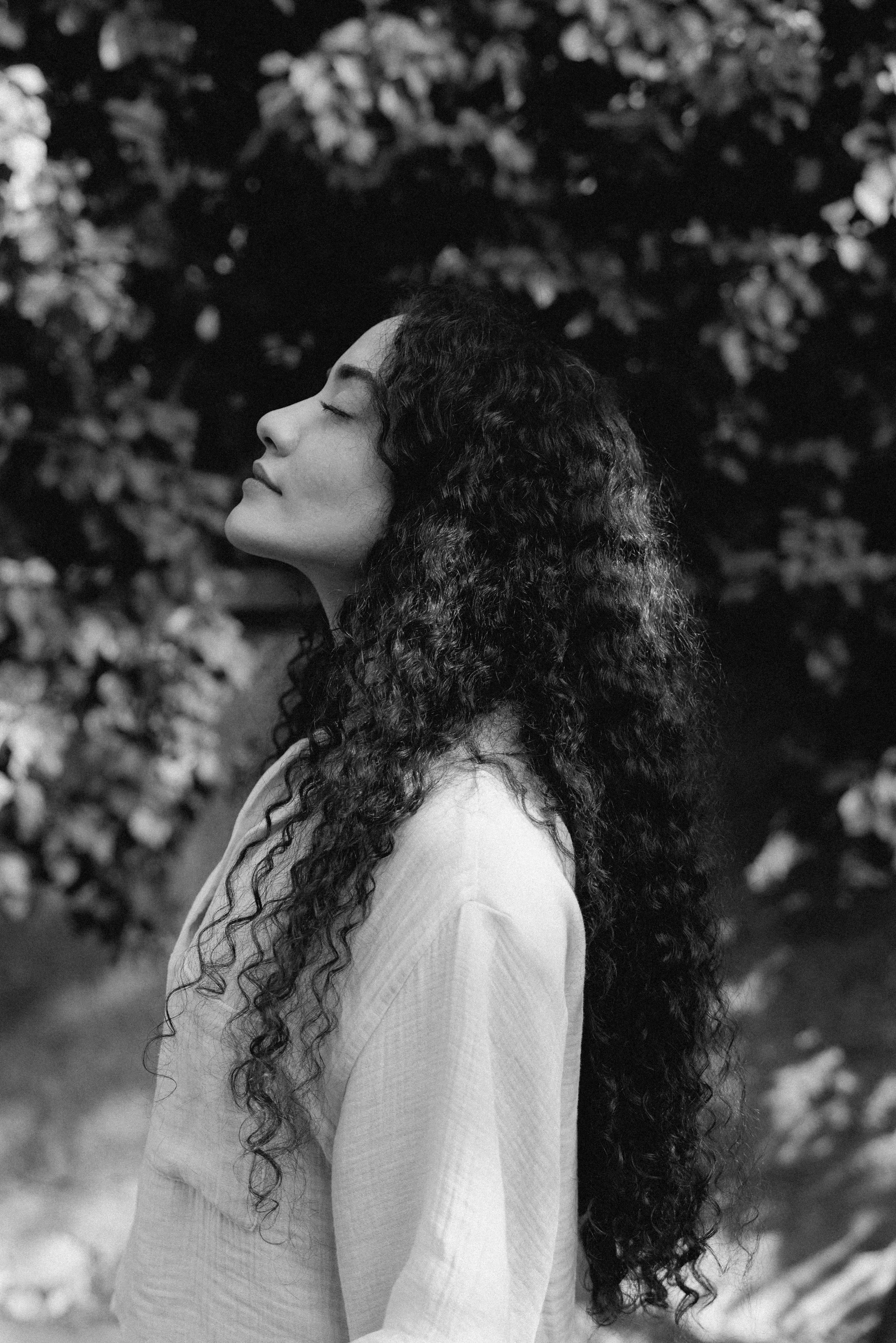 A woman with long curly hair standing outdoors with her eyes closed, wearing a light-colored top, and surrounded by blurred foliage in the background.