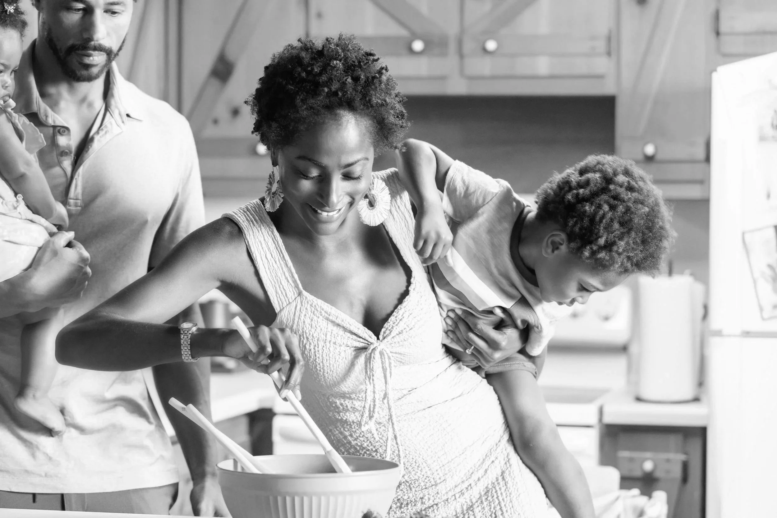 Black and white photo of a woman with short curly hair smiling as she cooks in the kitchen .
