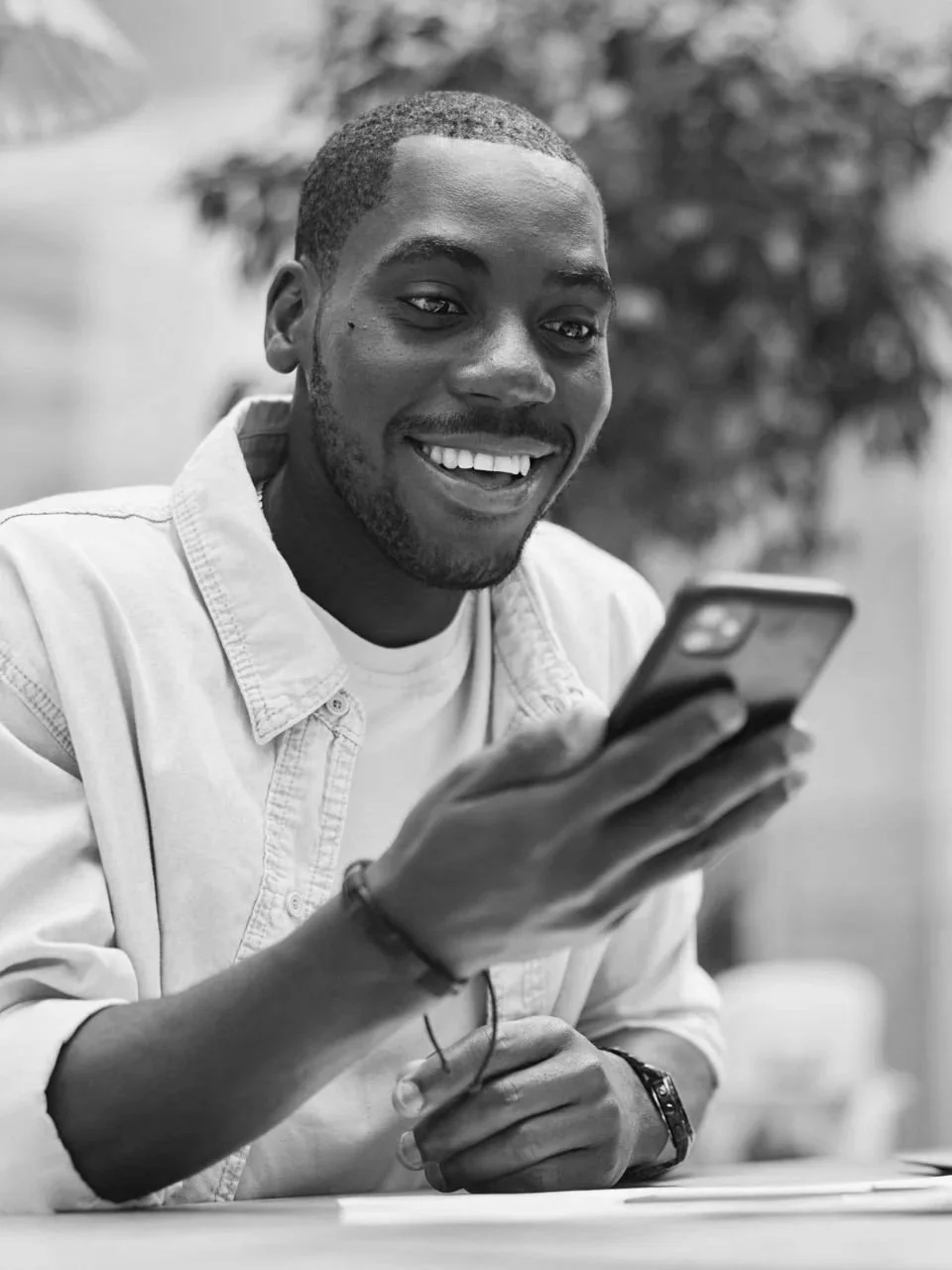 A smiling young man with a short beard and mustache, wearing a light-colored denim shirt, looking at his smartphone in an outdoor setting.