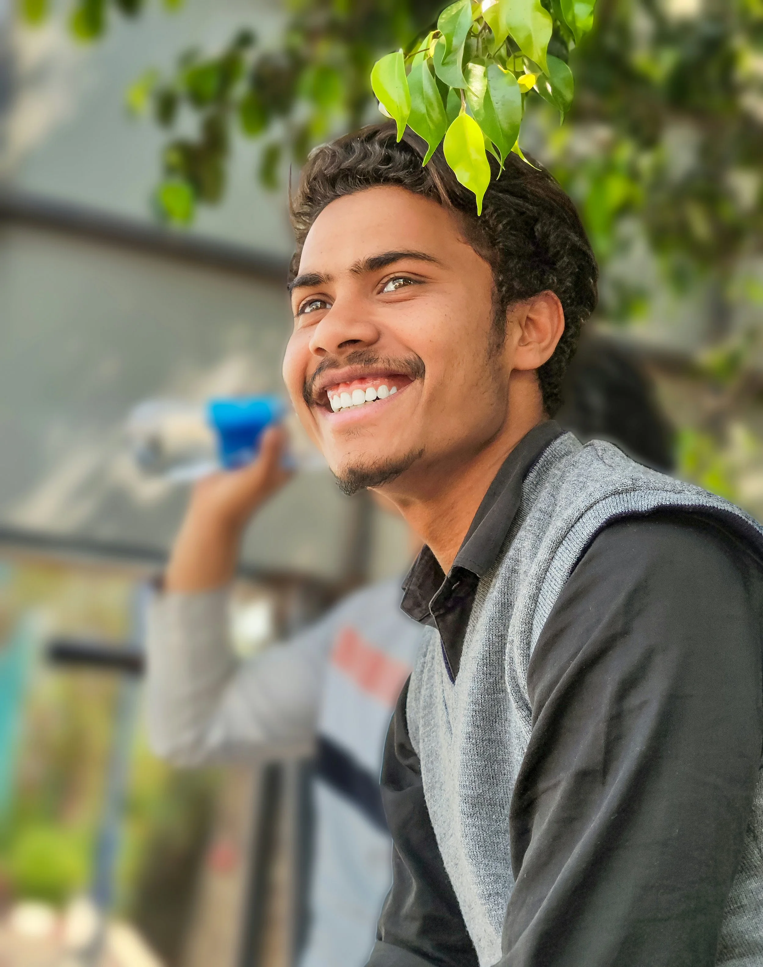 Close-up of a smiling young man with curly hair, wearing a gray and black shirt, outdoors with green leaves overhead and blurred background.