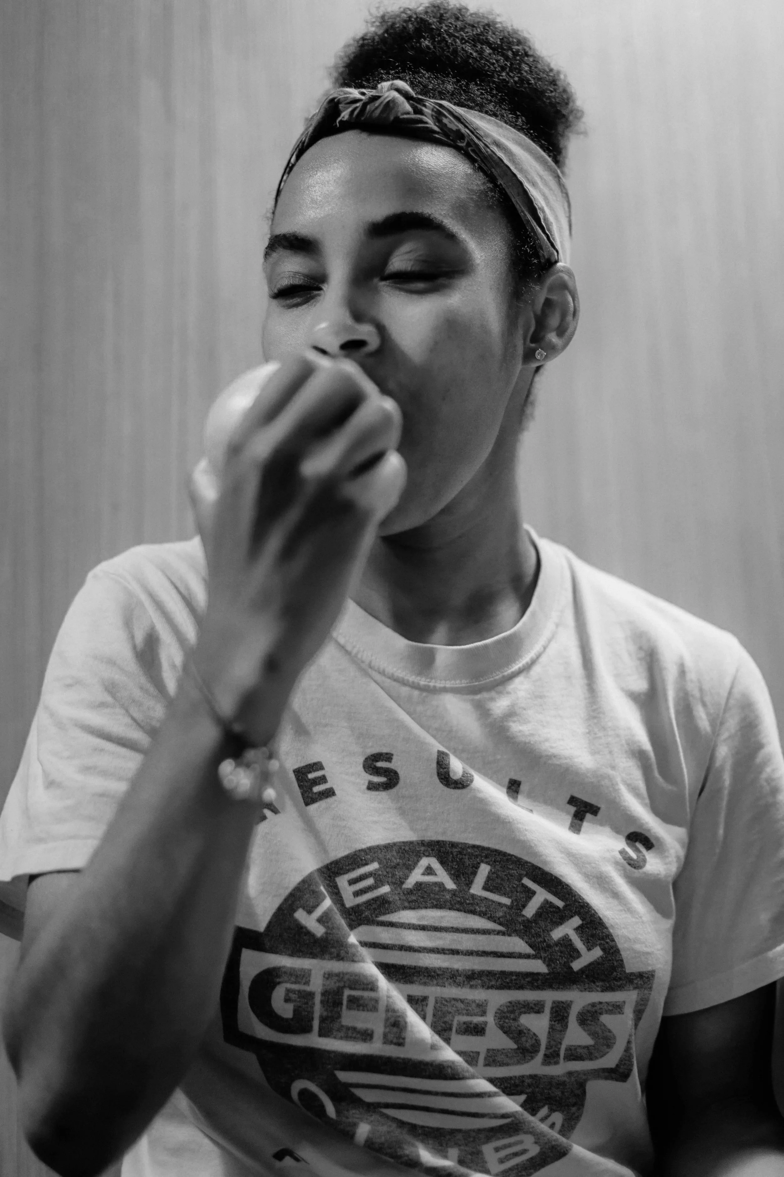 A woman with curly hair tied with a bandana, wearing a T-shirt, is smiling and holding her hand near her face.