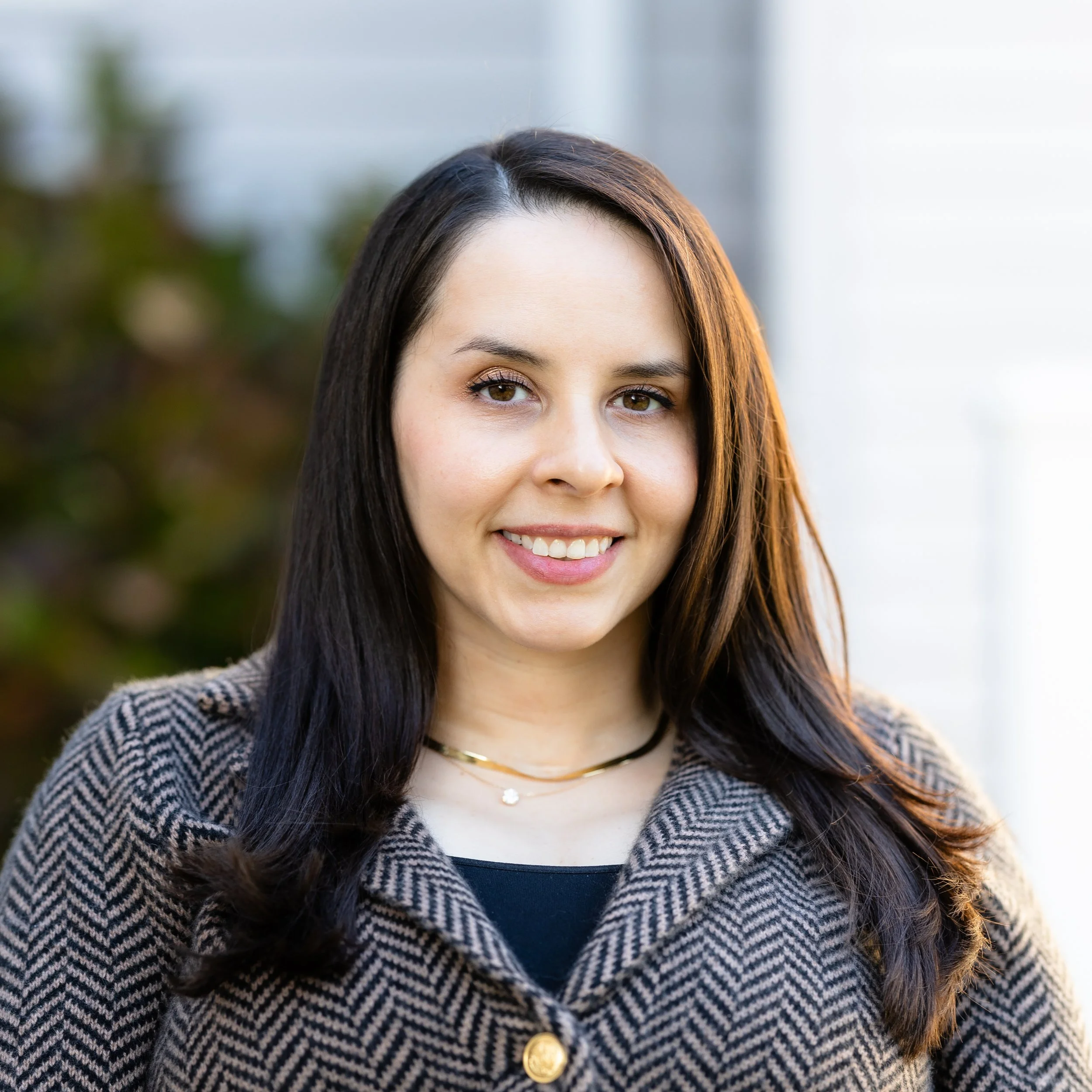 Tierney Cauley, owner of Eunoia Mental Health and Wellness in NJ, wearing a black top and a checkered coat with gold buttons, outdoors with a blurred background.