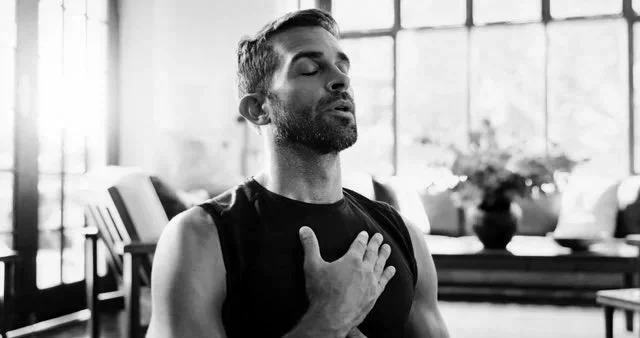 A man with a beard and closed eyes, touching his chest, in a room with large windows and potted plants, appearing to be meditating or in deep thought.