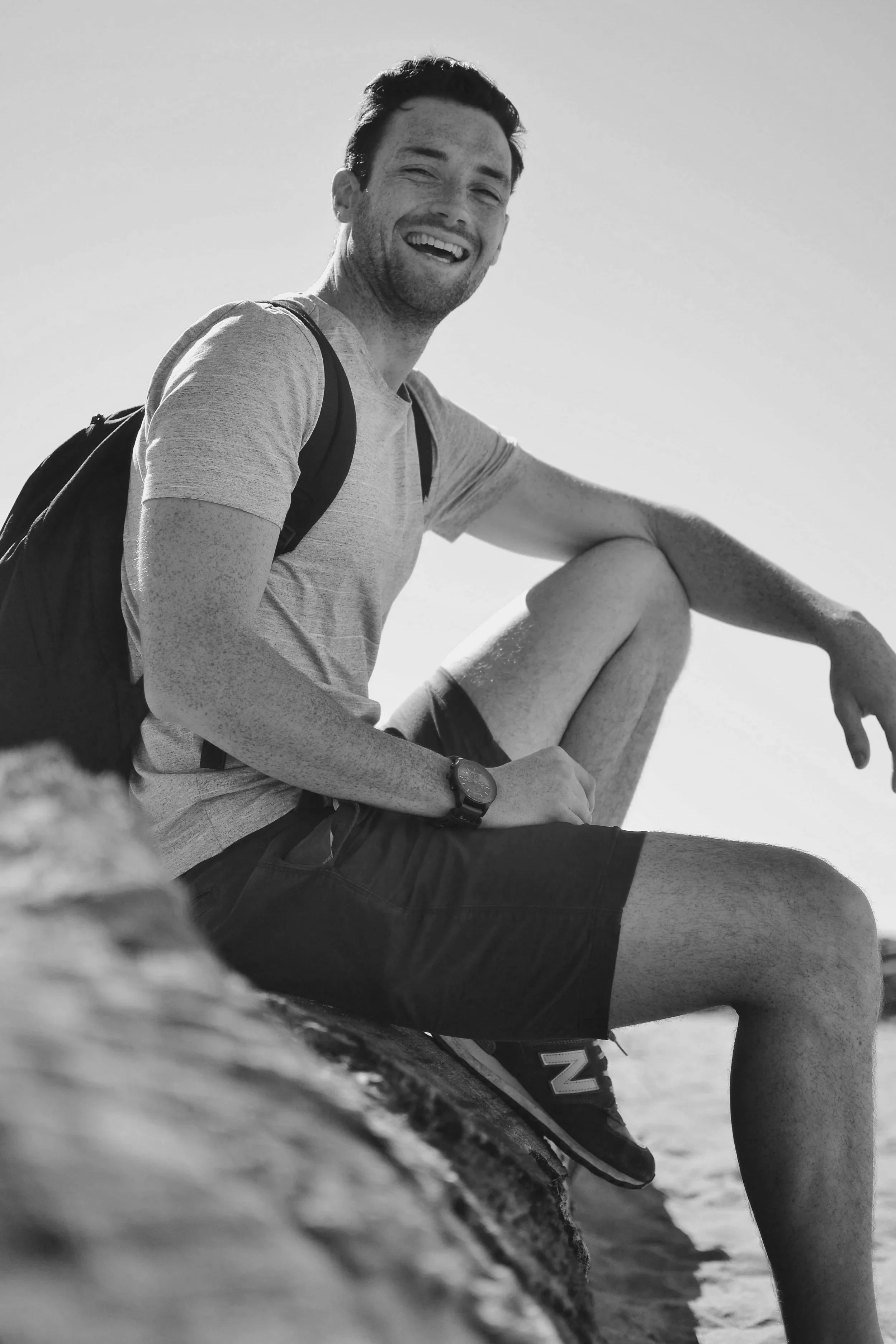 Black and white photo of a young man with dark hair, smiling, wearing a light-colored t-shirt and a backpack, sitting on a rock or large object outdoors with a clear sky in the background.