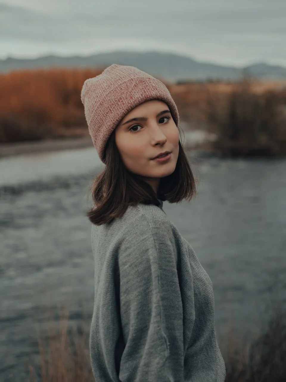A young woman with dark brown hair wearing a pink beanie and gray sweater standing outdoors near a body of water with trees and hills in the background.