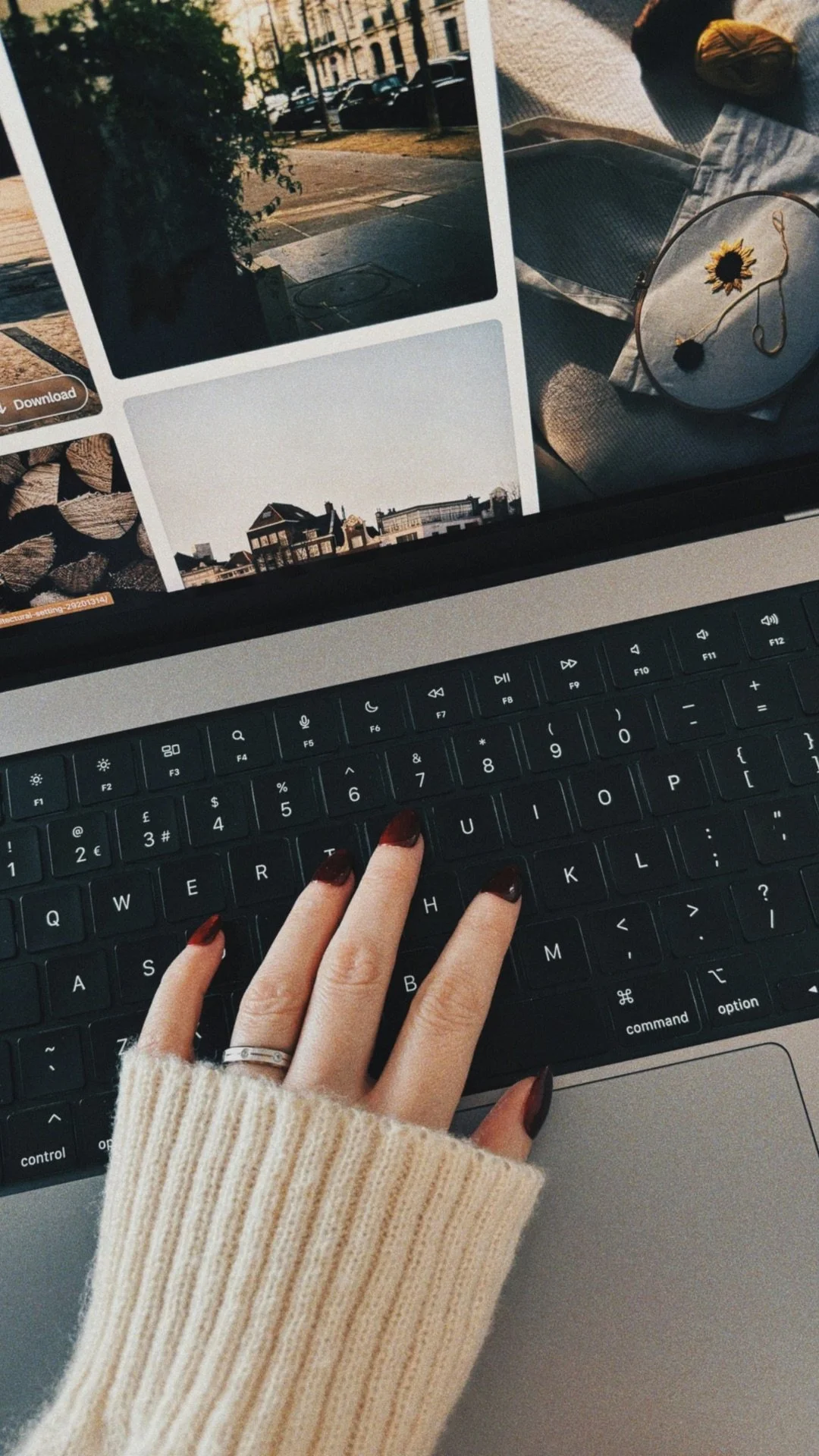 A person's hand with dark red nail polish on a keyboard, with a sweater sleeve visible. On the screen, there are several photos of rooftops, a street scene, and outdoor scenes.