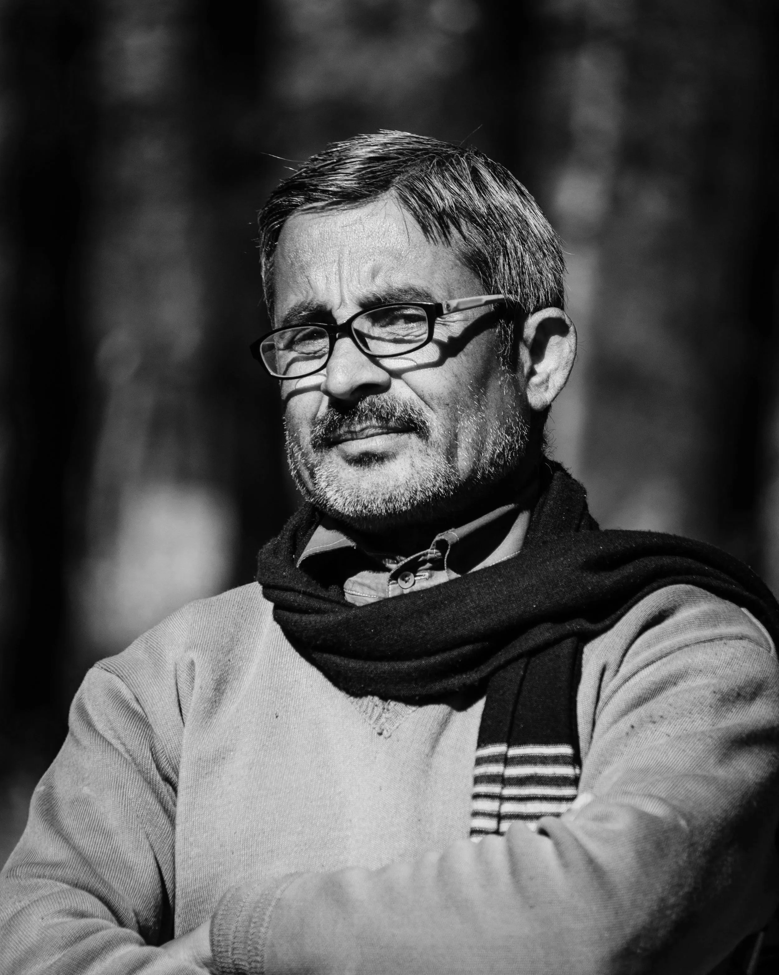 A black and white photo of a middle-aged man with glasses, short dark hair, and a beard, seated outdoors with his arms crossed. He is wearing a light-colored sweater and a scarf.