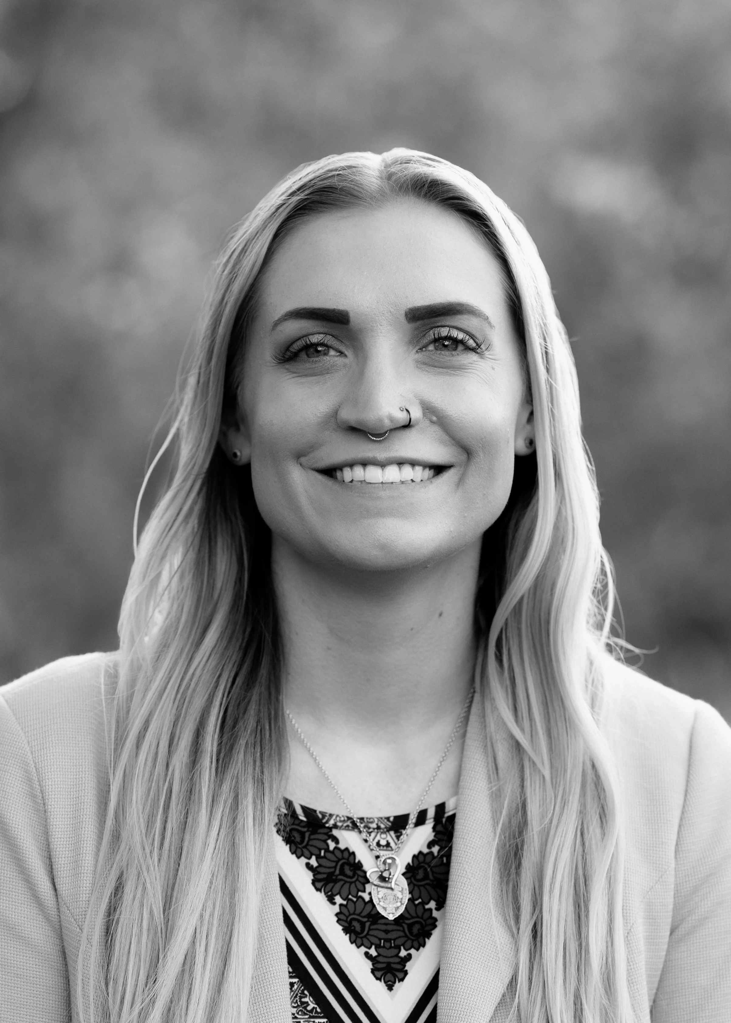 Haley Clancy, a therapist at Eunoia Mental Health and Wellness in NJ, with long hair, wearing a necklace, a patterned top, a light-colored blazer, and hoop earrings, standing outdoors against a blurred background.