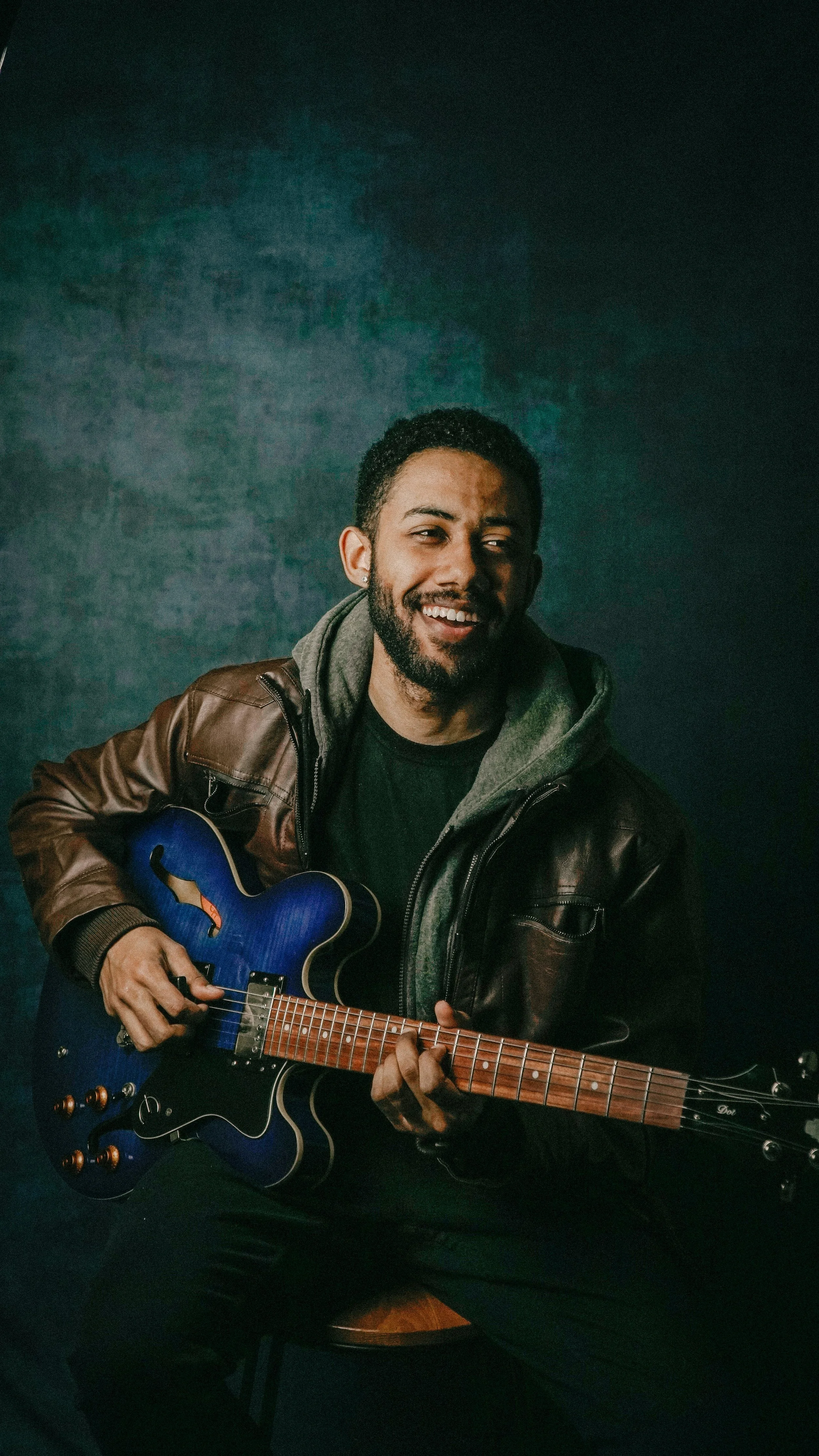 A man playing a blue electric guitar, sitting against a dark textured background, wearing a brown leather jacket and a gray hoodie, smiling.