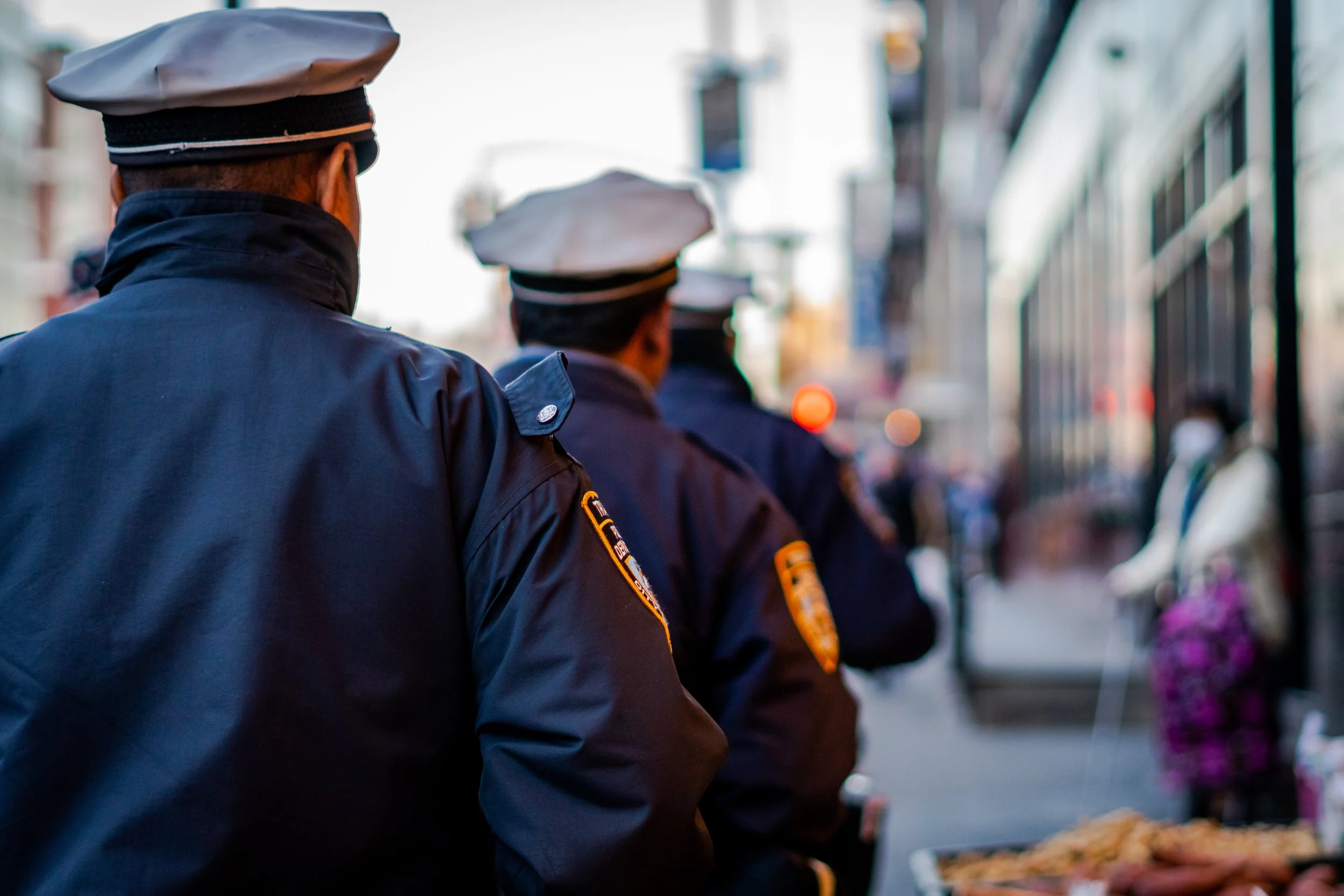 A group of police officers standing in a line outdoors, facing away from the camera, with city buildings in the background.