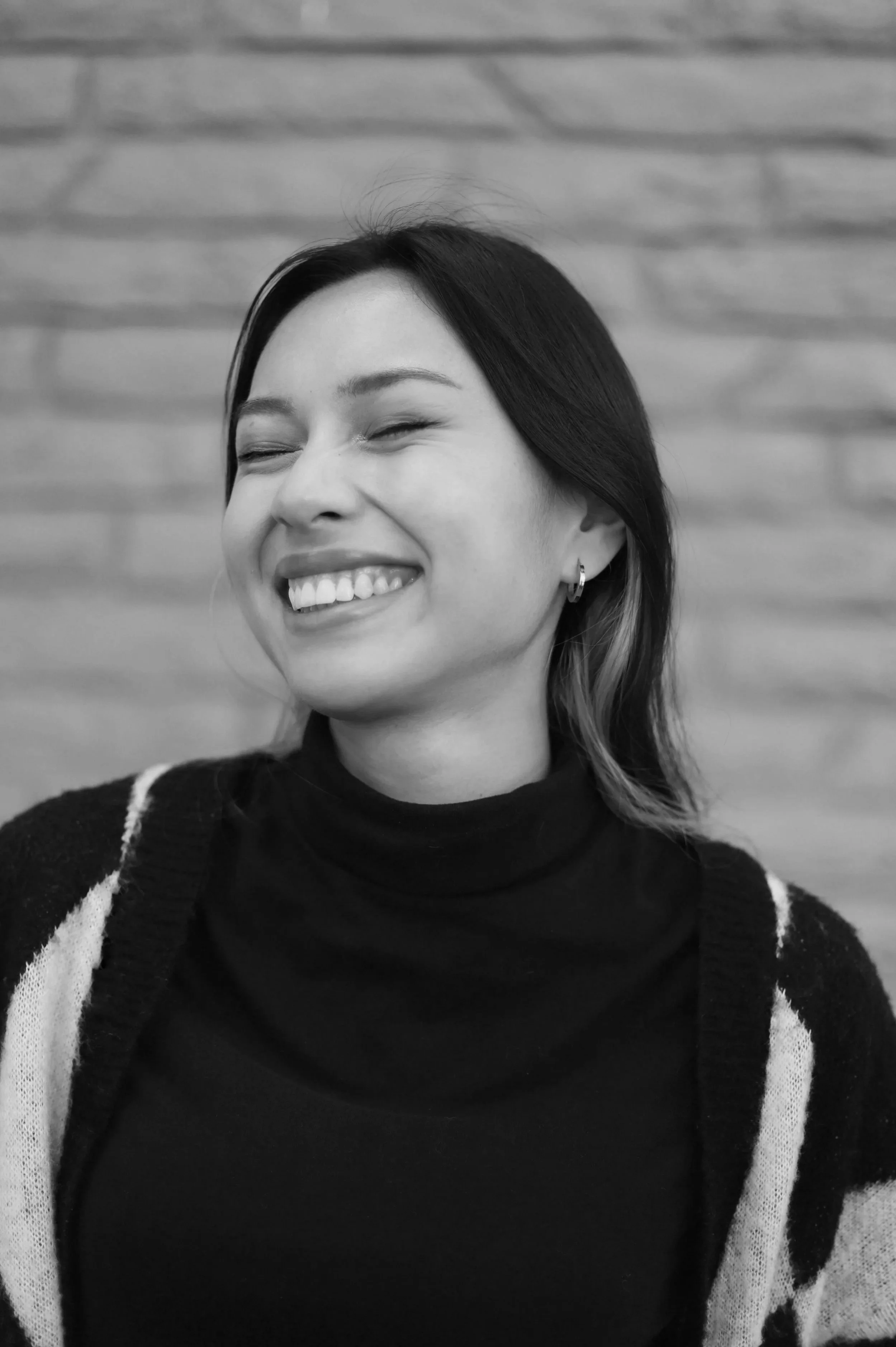 A woman with black hair, smiling with closed eyes, wearing earrings, a black turtleneck, and a black-and-white patterned cardigan, standing in front of a wooden wall.