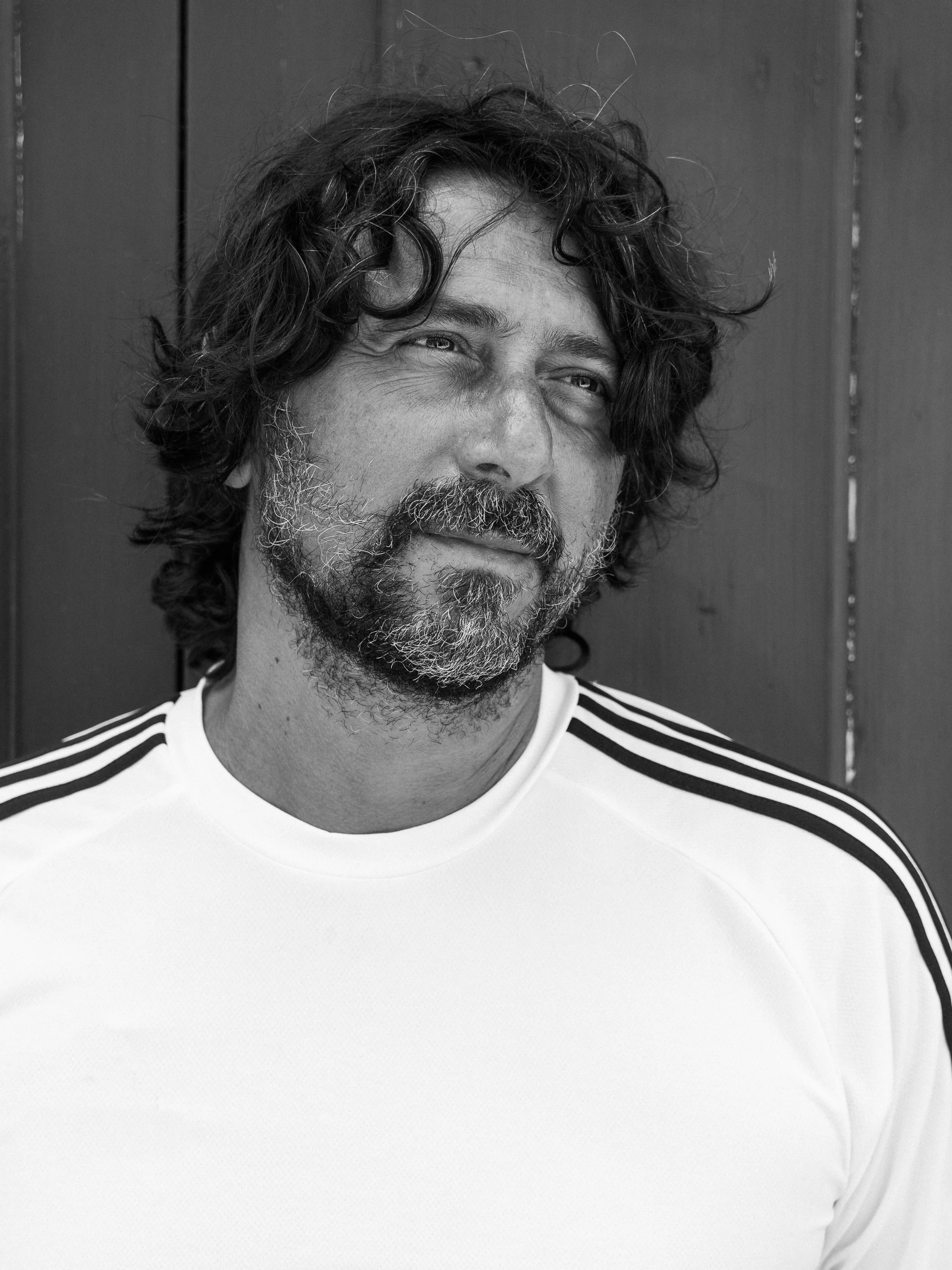 A black and white photo of a man with curly hair and a beard, wearing a white shirt with black stripes on the shoulders, standing in front of a wooden wall.