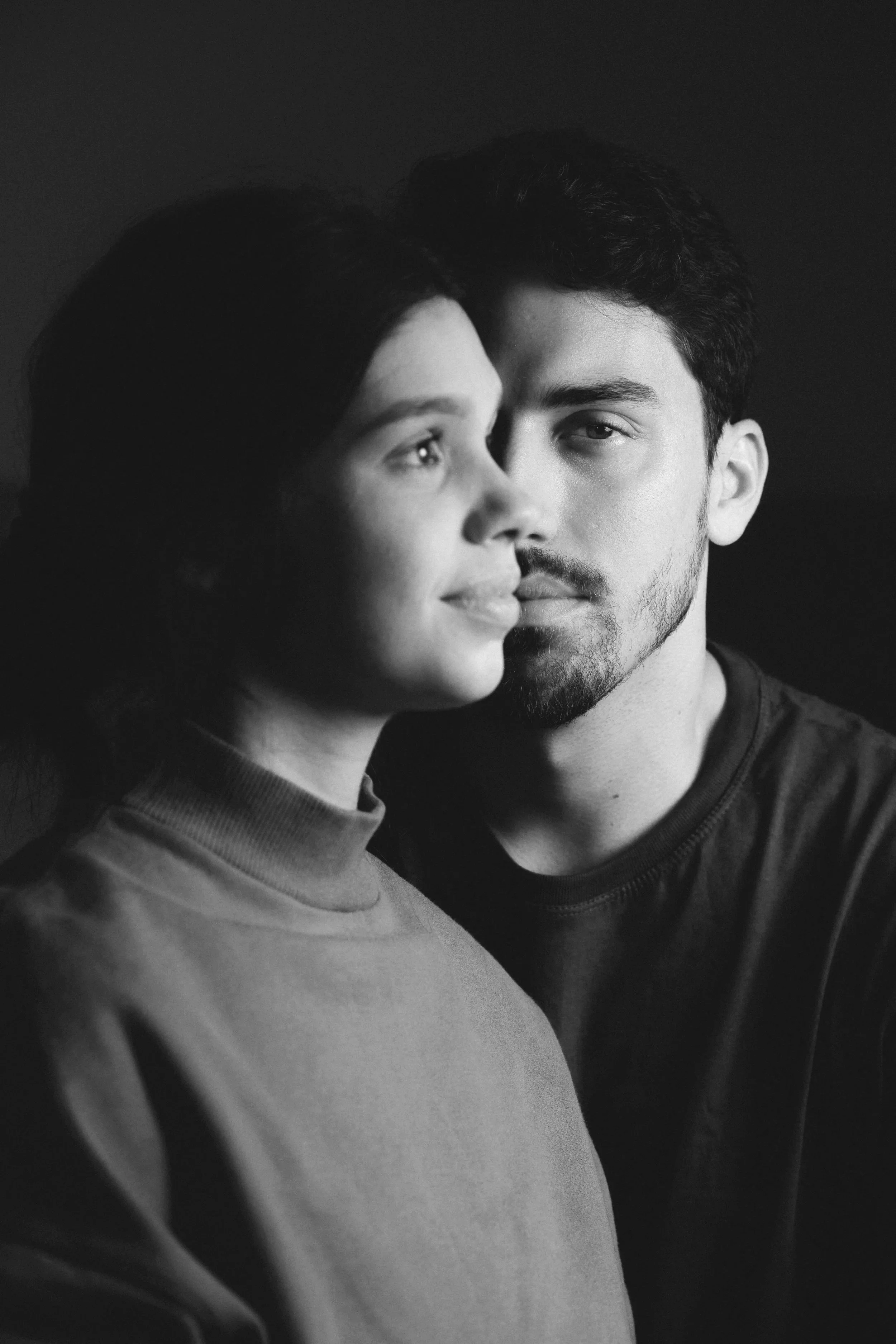 Black and white photo of a young man and young woman standing close together, both looking at the camera with neutral expressions. The woman is in the foreground, slightly to the left, and the man is behind her, slightly to the right.