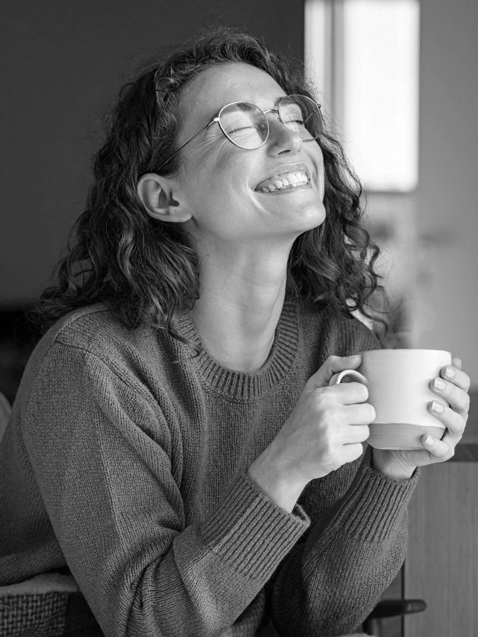 A woman with curly hair and glasses smiling with her eyes closed, holding a mug, seated indoors near a window.