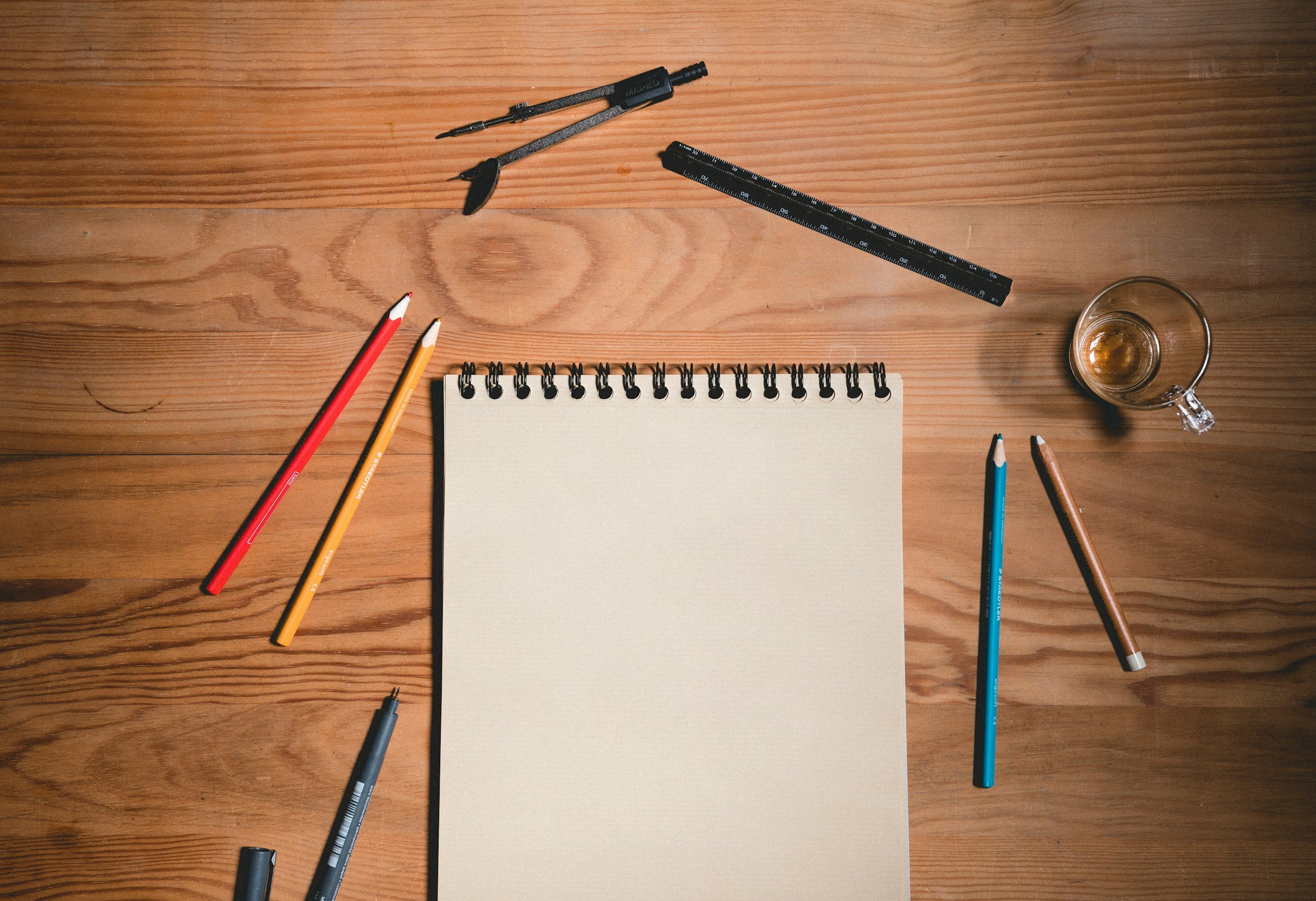 Wooden desk with drawing tools, colored pencils, a black pen, a mechanical pencil, a ruler, an empty spiral notebook, and a glass cup with a small amount of liquid.