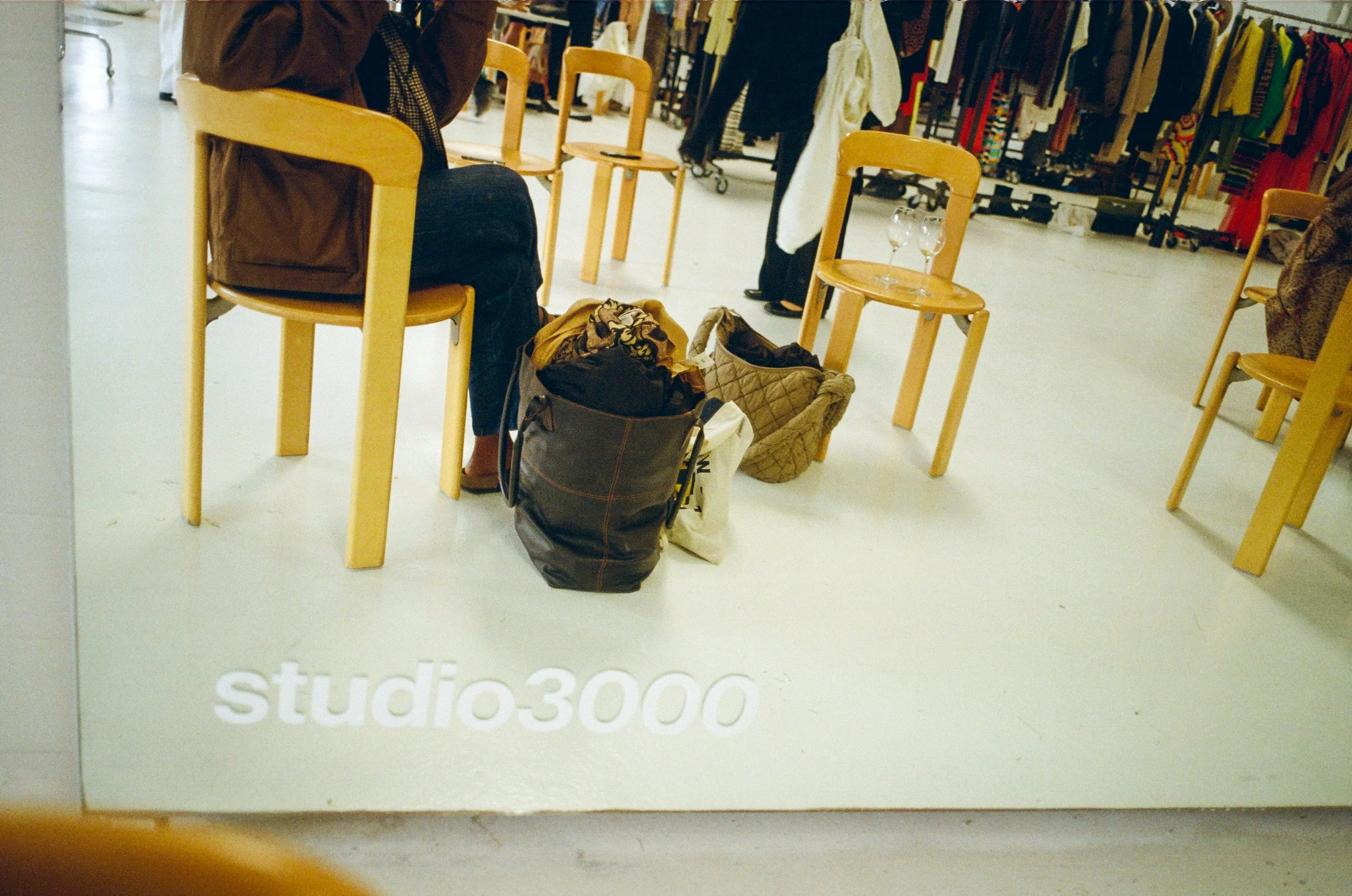 Miroir dans un magasin de vêtements, avec des chaises en bois, un sac à mains rempli de vêtements, et quelques vêtements suspendus en arrière-plan.