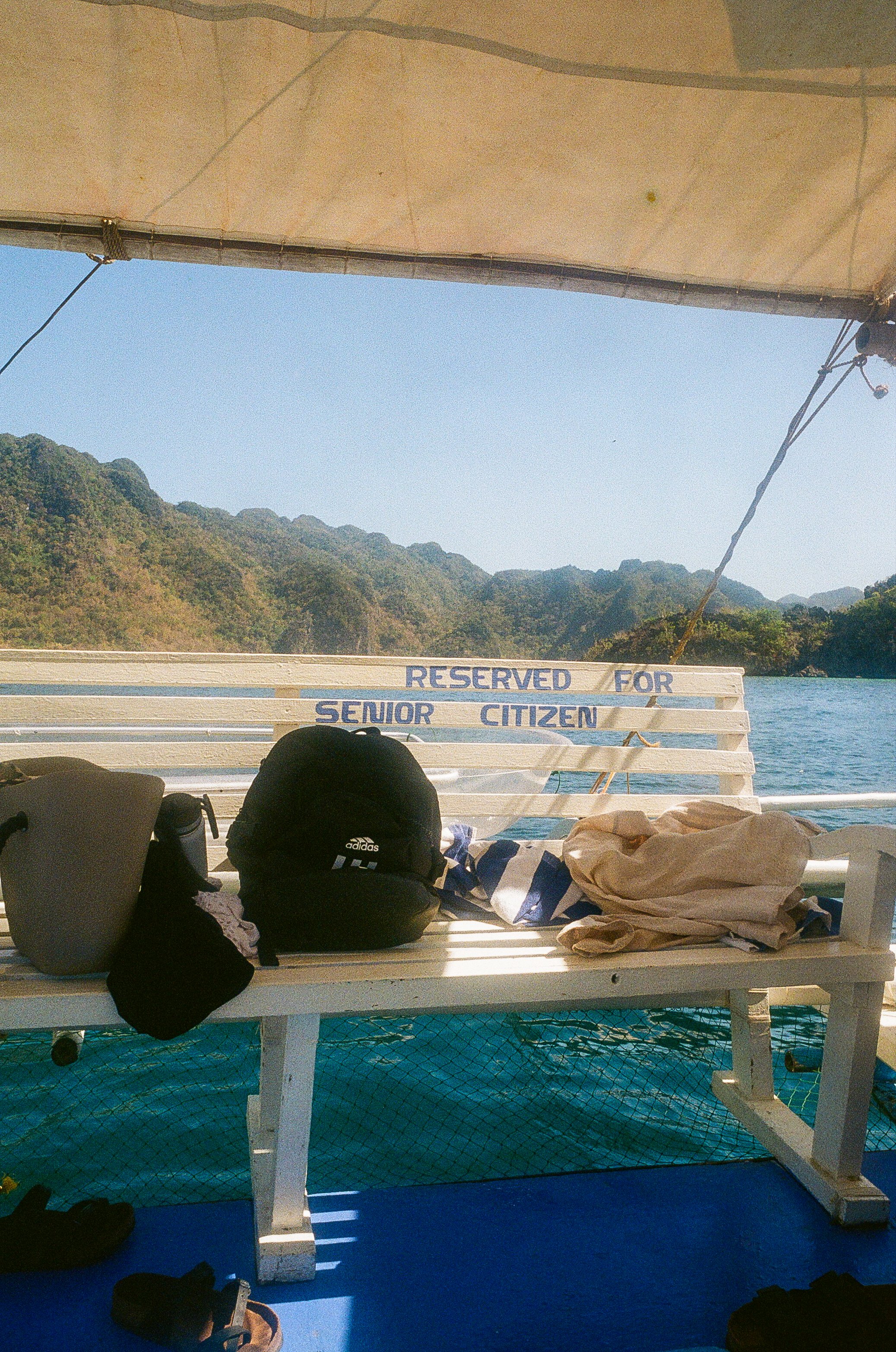 Banc réservé aux personnes âgées sur un bateau avec des montagnes en arrière-plan, des sacs et vêtements posés dessus, et une vue sur l'eau.