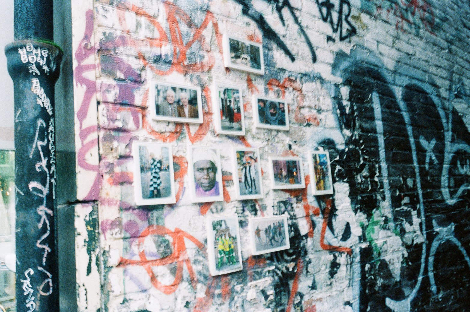 Mur de briques recouvert de graffiti avec plusieurs cadres de photos accrochés, incluant des portraits et scènes de rue, dans un style urbain.