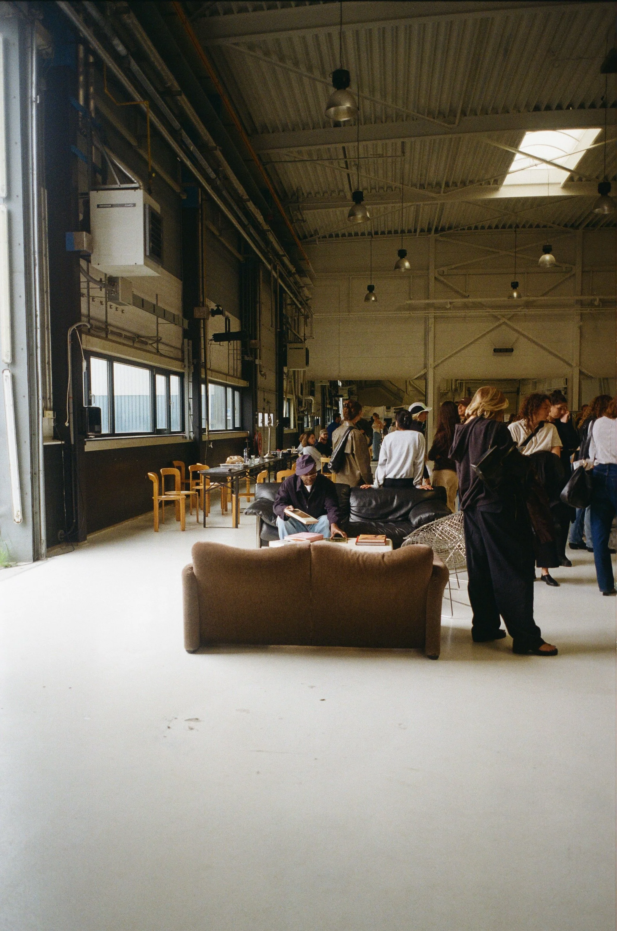 Un groupe de personnes à l'intérieur d'un espace industriel ou d'un hangar, avec des meubles et des fenêtres, certaines personnes sont assises et d'autres discutent ou circulent.