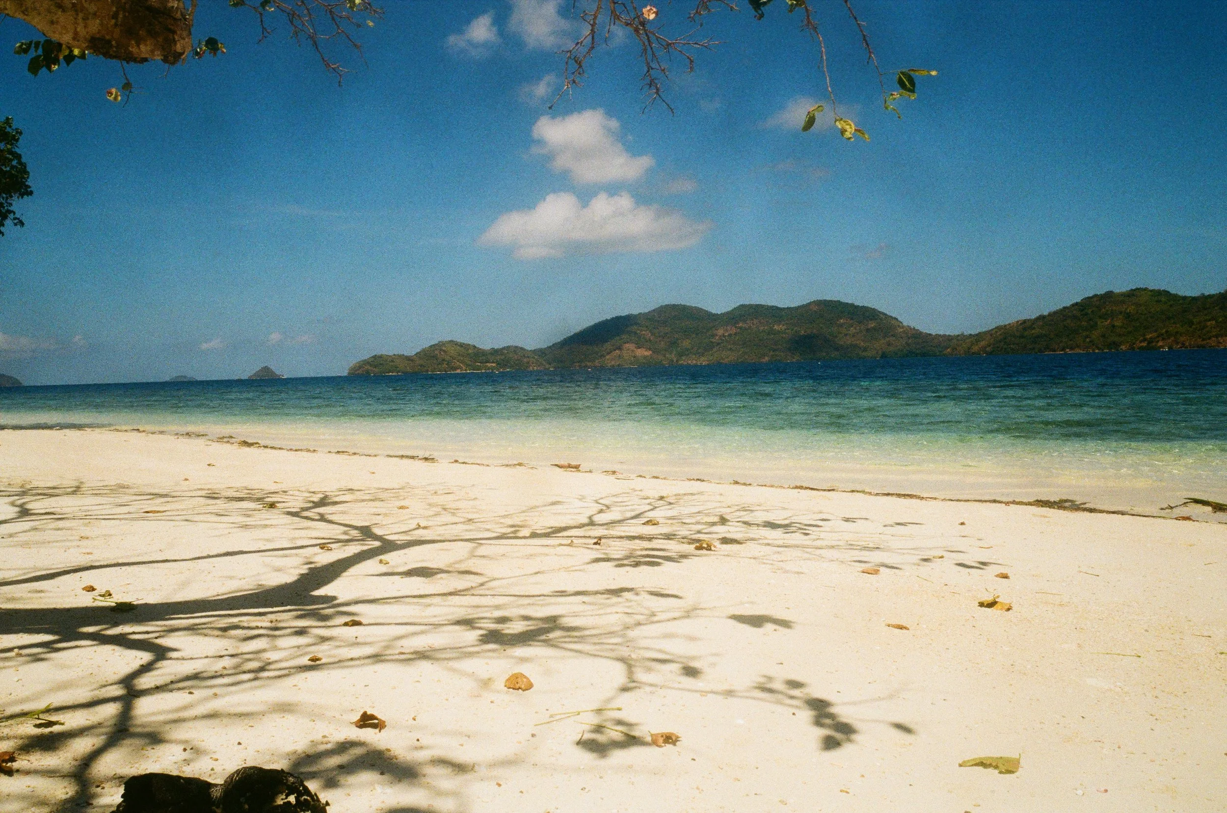 Plage de sable blanc avec des arbres et leurs ombres, mer bleue claire, montagnes en arrière-plan, ciel bleu avec quelques nuages.