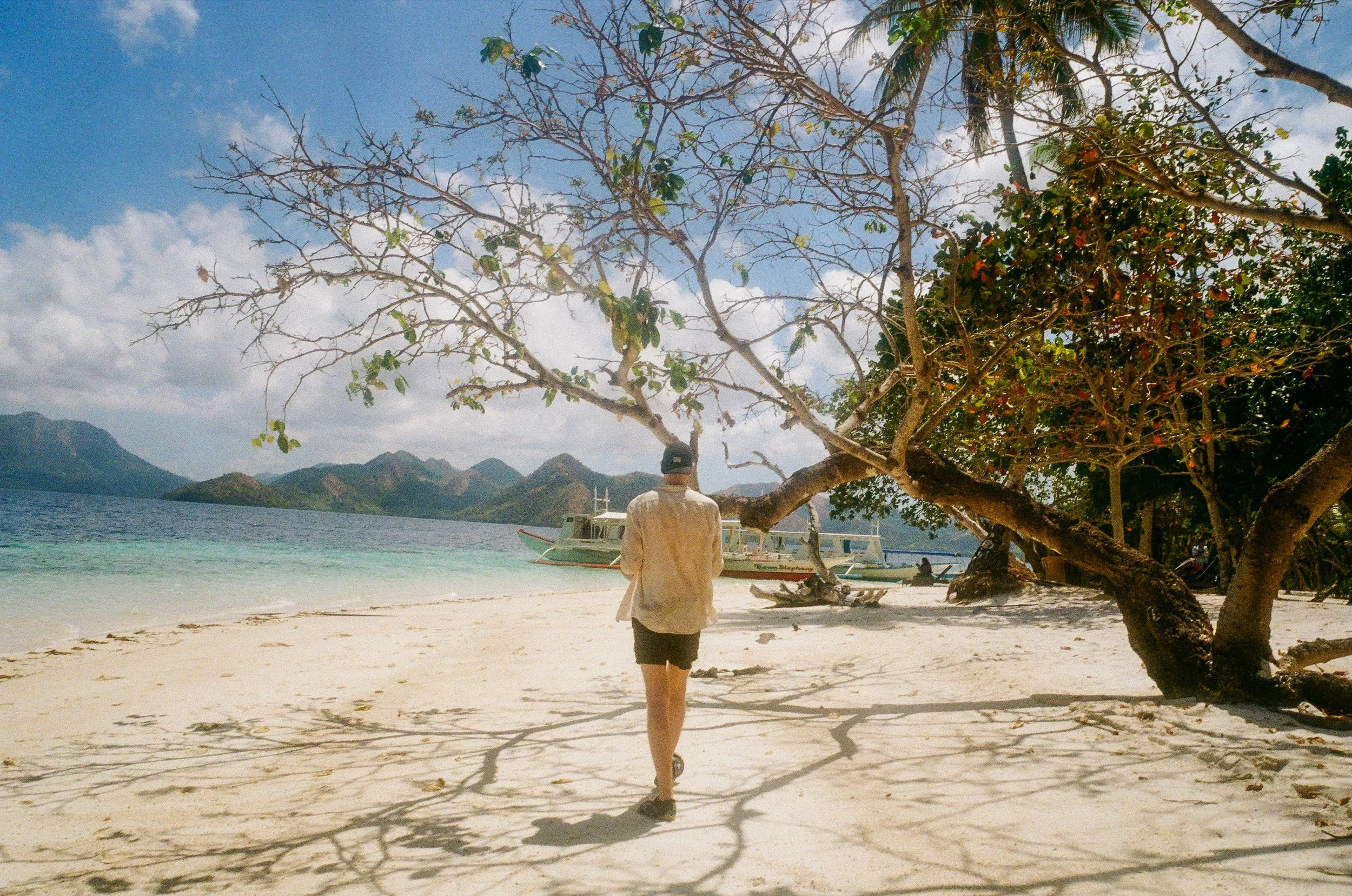 Personne marchant sur une plage de sable blanc avec des branches d'un arbre étendu sur le sol, vue sur la mer, des montagnes en arrière-plan, et un bateau à moteur dans l'eau.