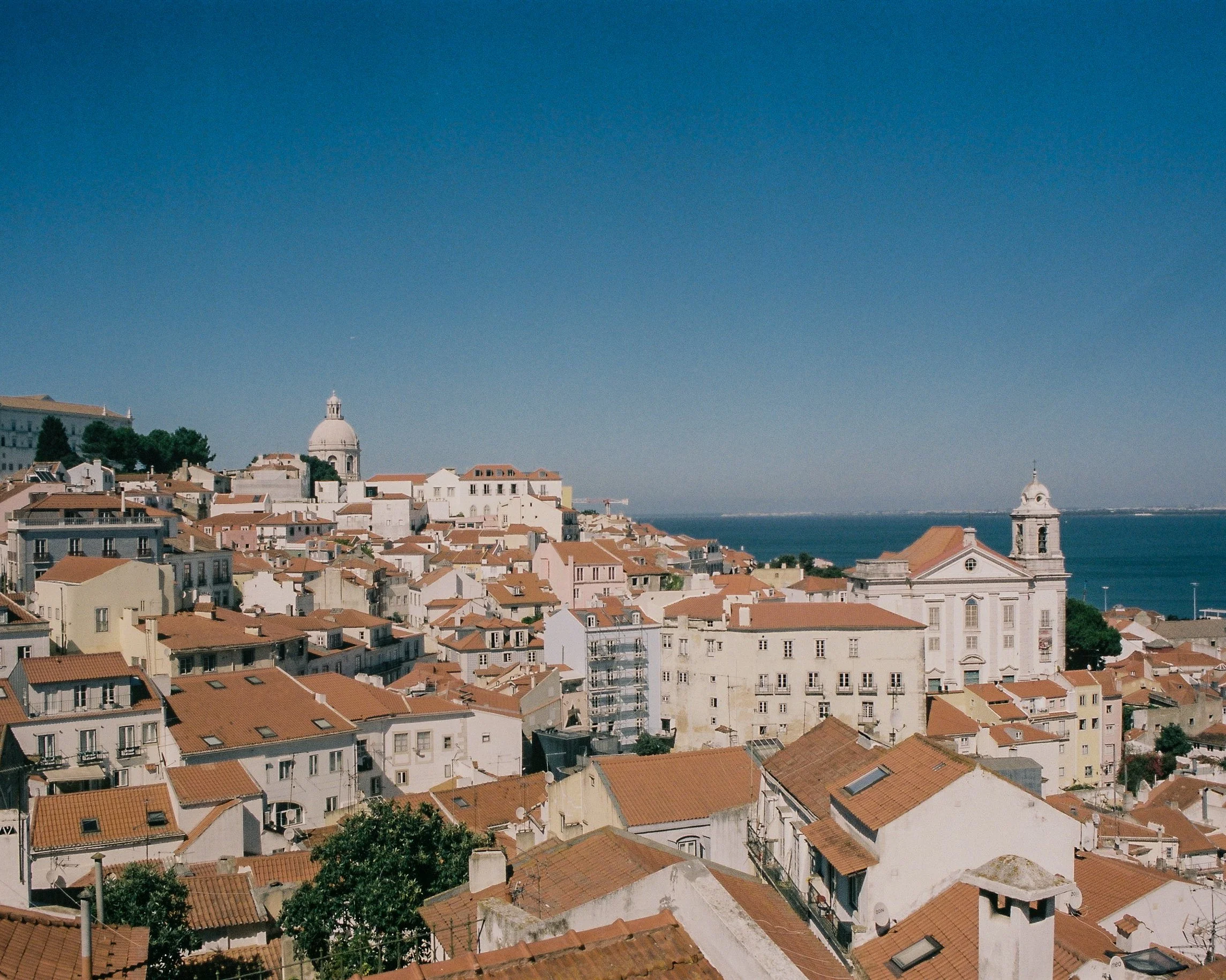 Vue aérienne d'une ville côtière avec des bâtiments aux toits en tuiles rouges, une grande église blanche avec un clocher, et la mer en arrière-plan sous un ciel bleu clair.