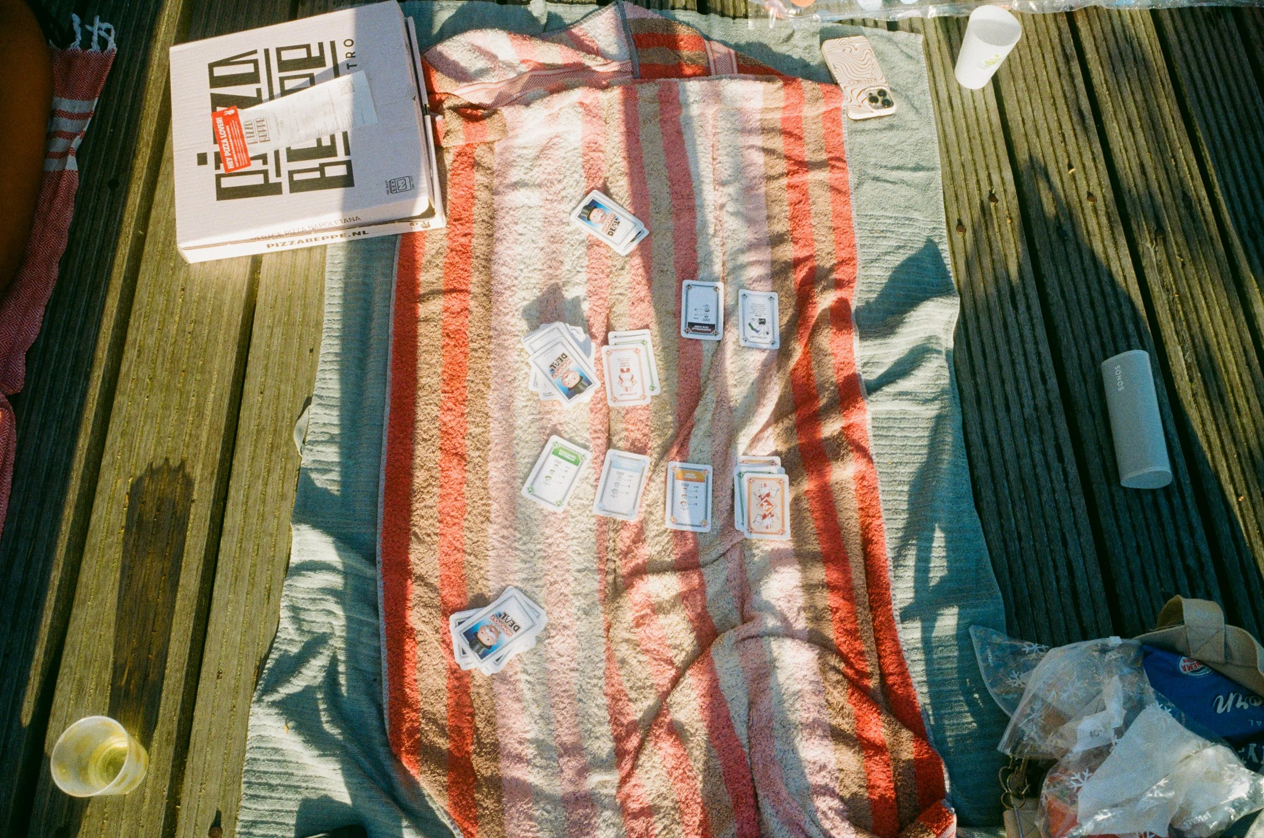 Jeu de société de cartes avec une pizza à emporter, un verre en plastique blanc, un haut-parleur portable, et un sac de nourriture, le tout posé sur un tapis à rayures rouge et beige sur un pont en bois.