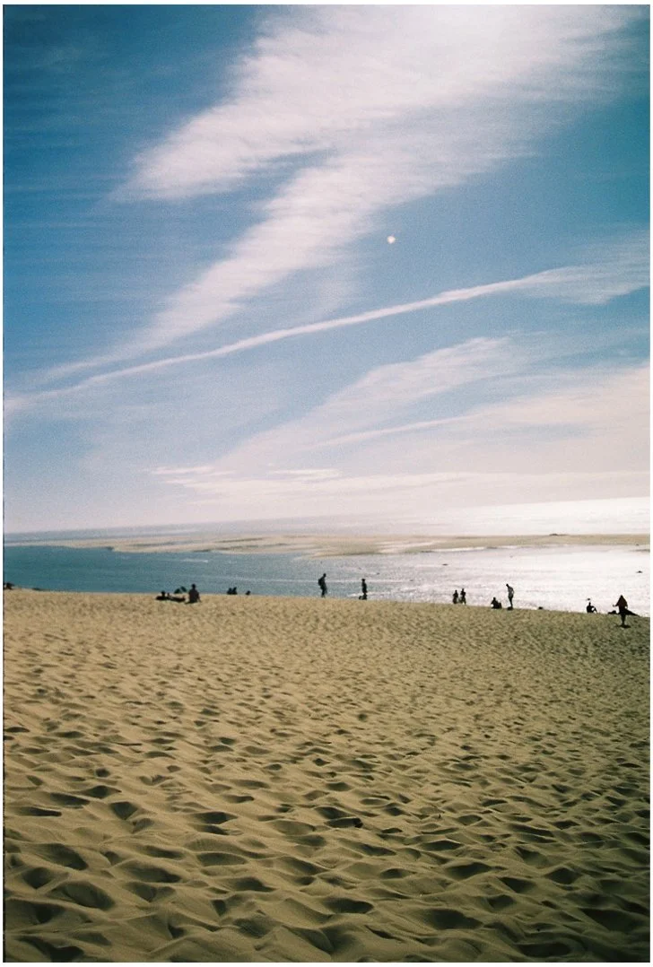 Plage de sable avec des personnes au bord de l'eau, ciel bleu avec quelques nuages et la lune visible
