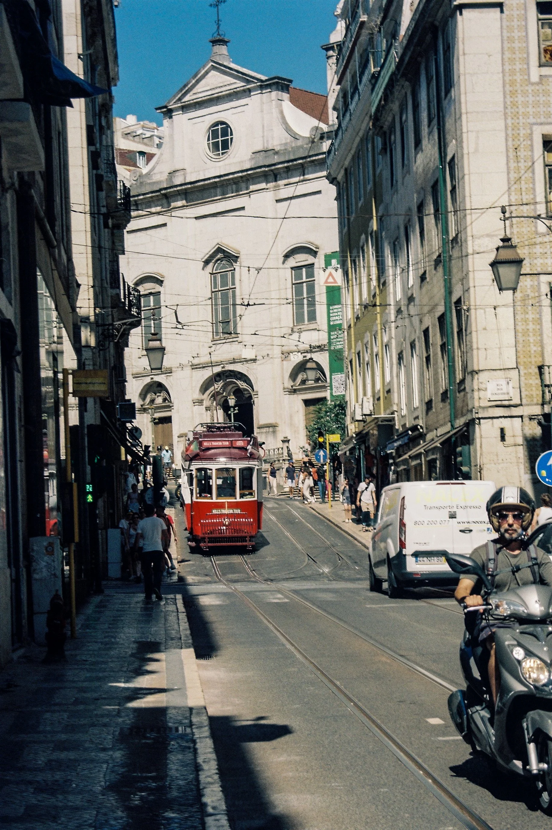 Rue en pente avec un tram rouge, une moto avec un homme portant un casque, des voitures blanches et des piétons sous un ciel bleu, devant une grande église blanche avec un toit en tuiles rouges à Lisbonne.