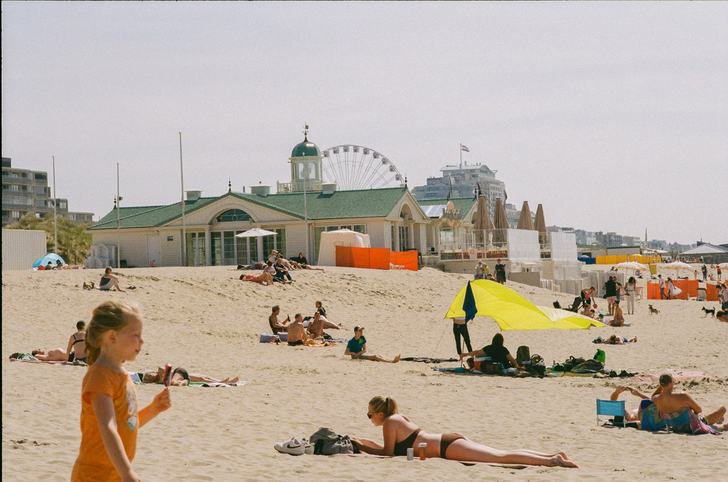 Plage avec des personnes allongées et assises sur le sable, parasol jaune, bâtiments en arrière-plan, grande roue en centre, ciel clair.