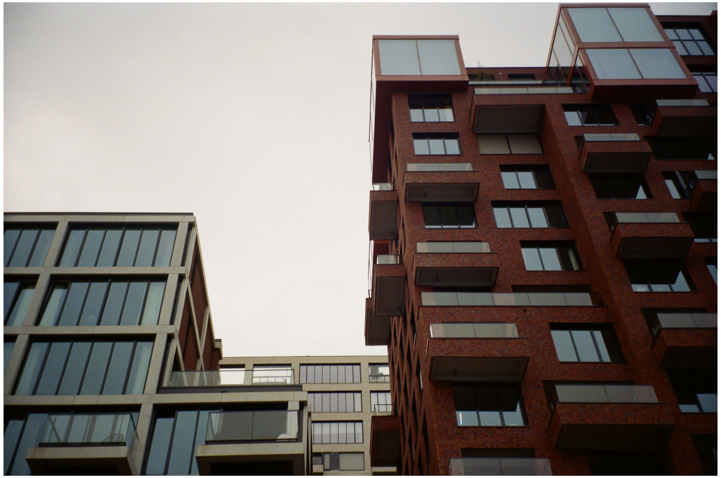 Deux immeubles modernes en contre-plongée, avec de nombreux balcons en saillie, en briques rouges et en verre, sous un ciel gris.