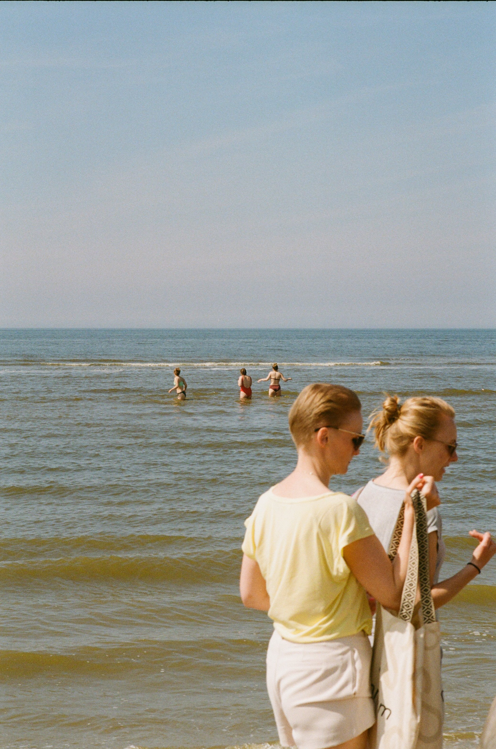 Deux femmes discutent sur la plage, avec trois personnes dans l'océan en arrière-plan et un ciel clair.