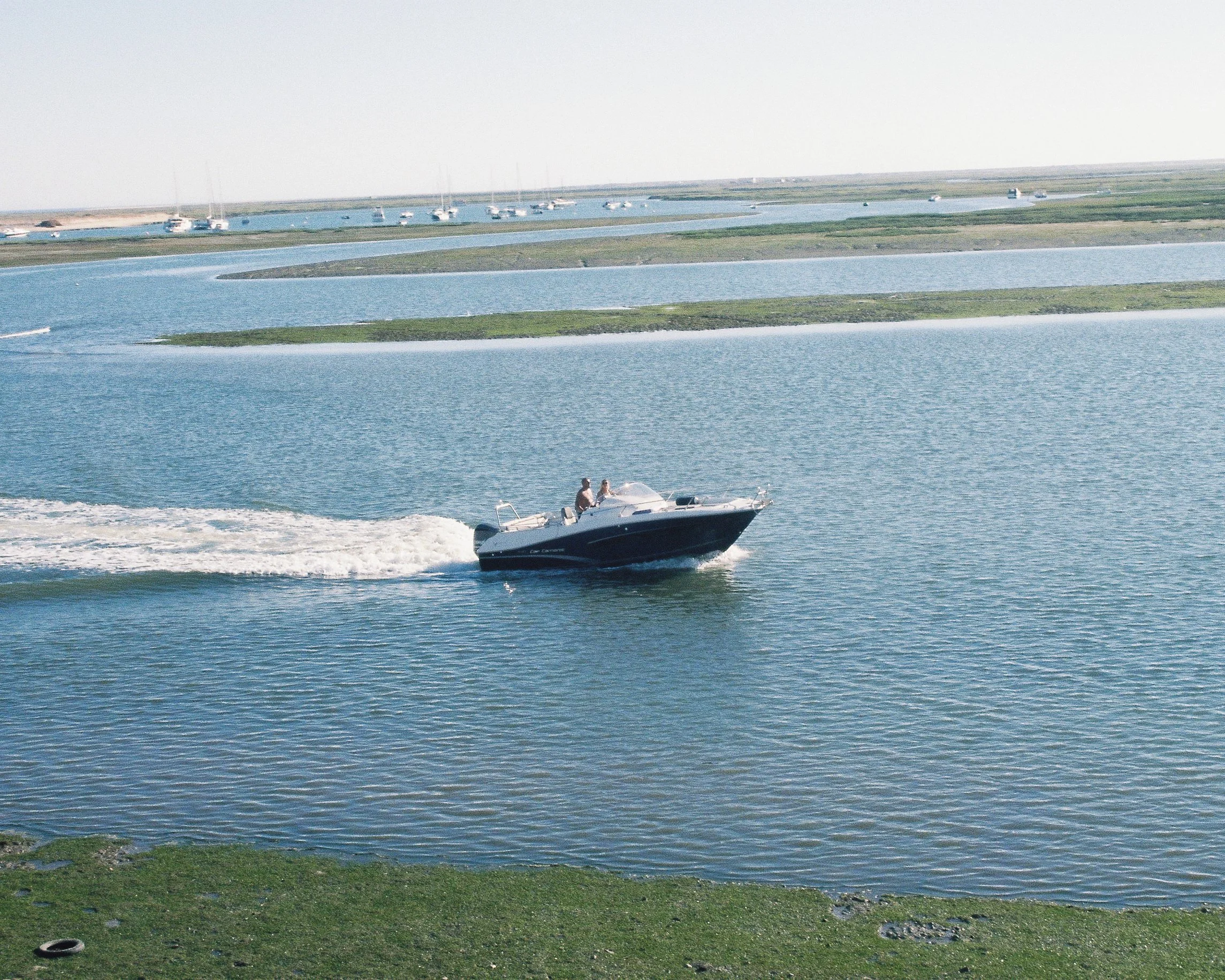Un bateau à moteur navigue sur une rivière ou un étang, avec plusieurs voiliers amarrés au loin et des îles ou bancs de sable recouverts d'herbe dans l'eau.