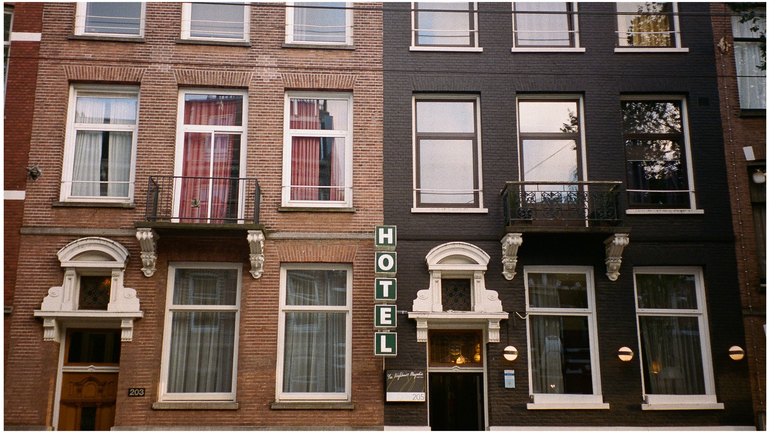 Façade d'un bâtiment avec un panneau vertical indiquant l'hôtel, comprenant des fenêtres à ferronnerie noire et des balcons, avec une partie en briques rouges et une autre en briques noires.